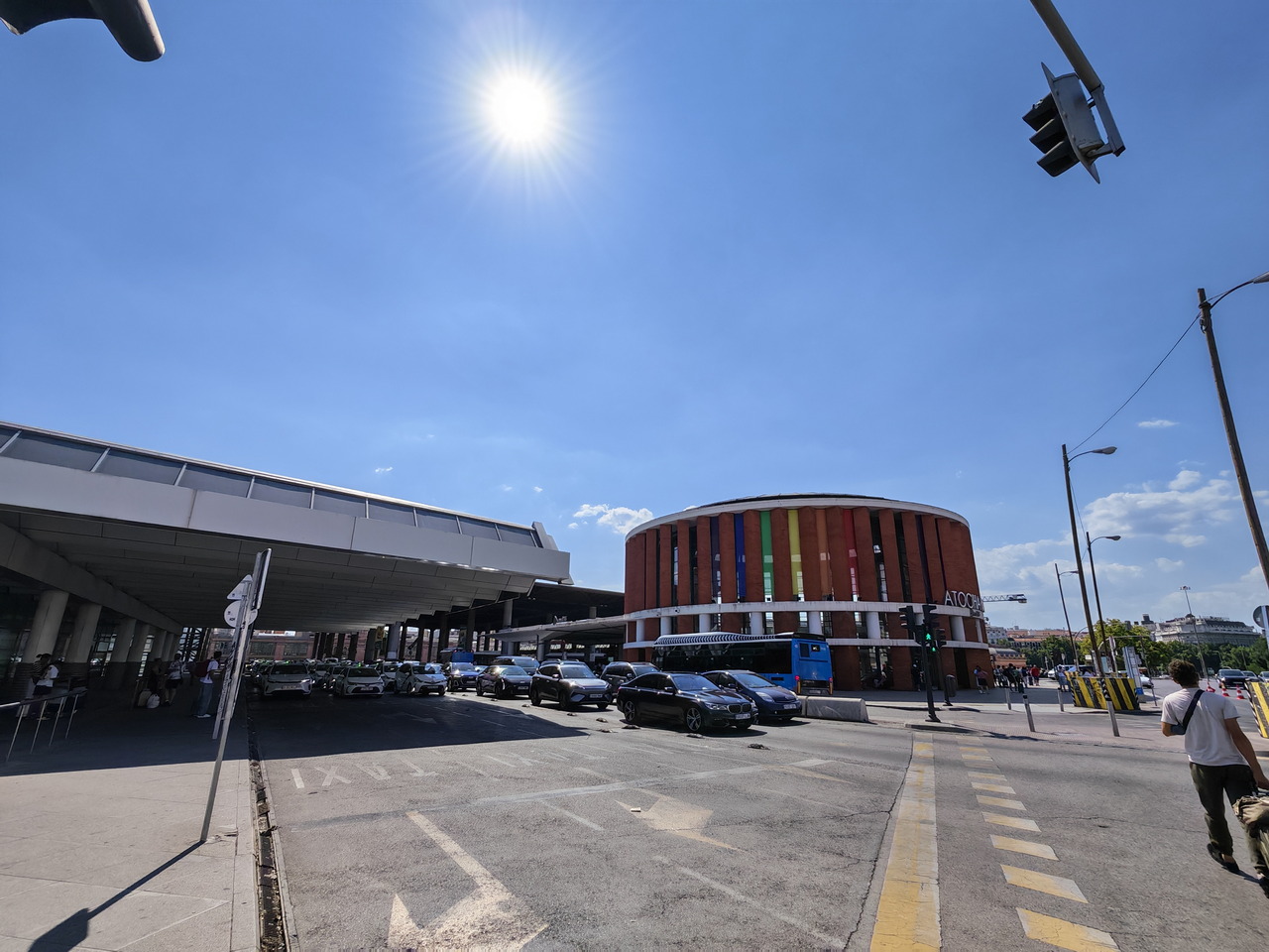 Estación de tren Atocha en Madrid con tráfico y cielo soleado. Edificio circular colorido al fondo.