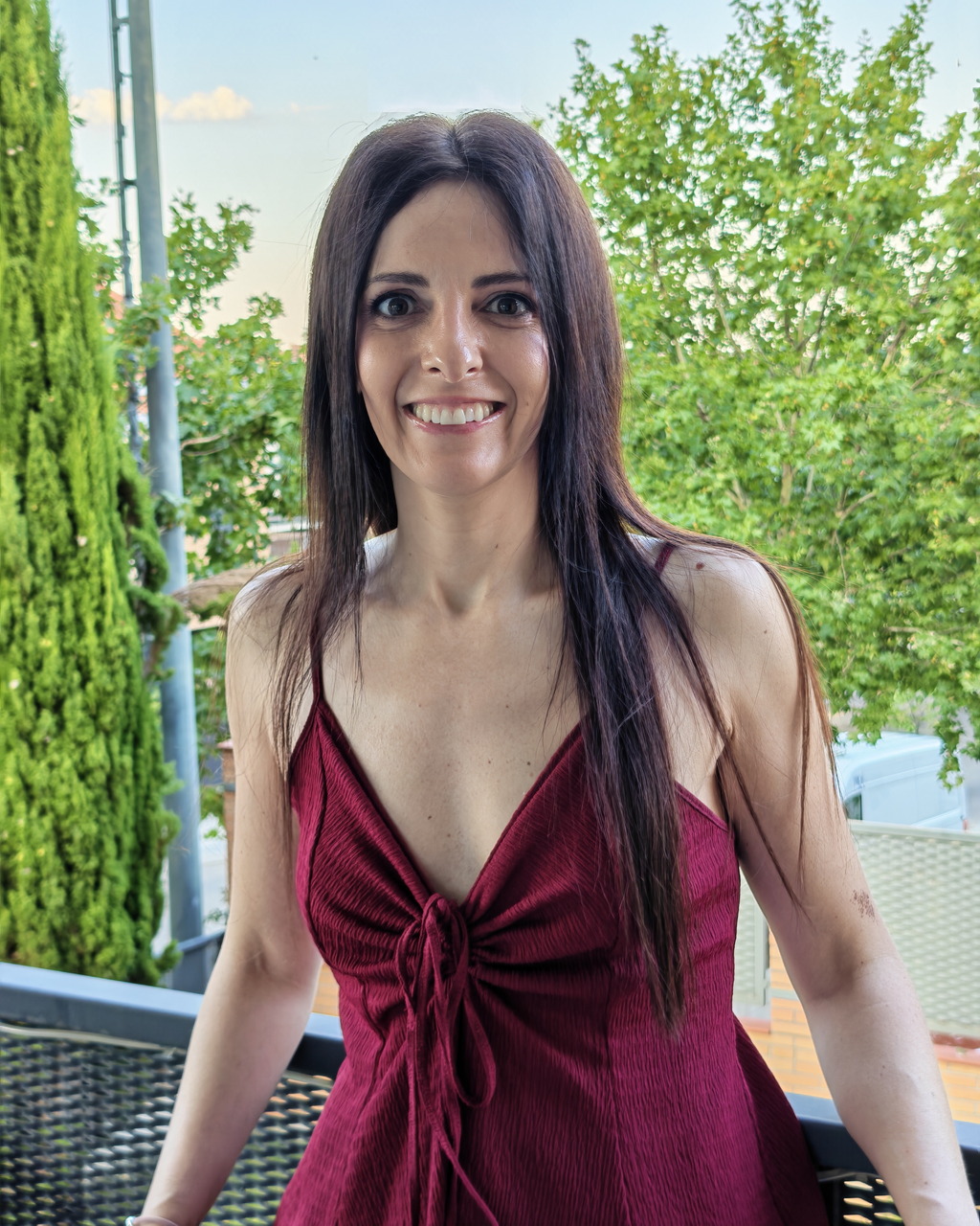 Mujer sonriendo con vestido rojo en un balcón, rodeada de árboles verdes en un día soleado.