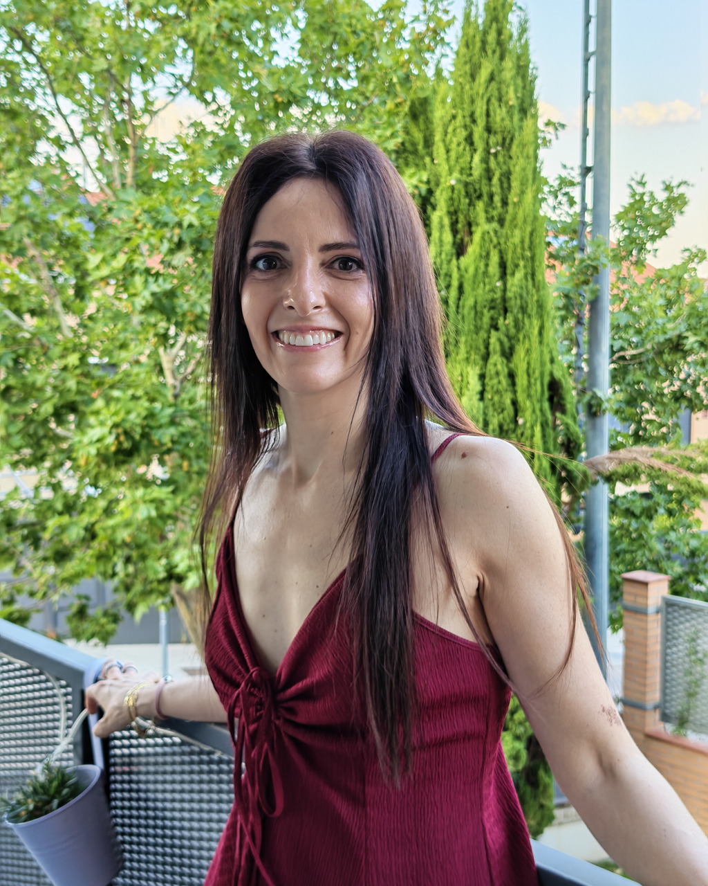 Mujer sonriente con vestido rojo en un balcón con fondo de árboles verdes.