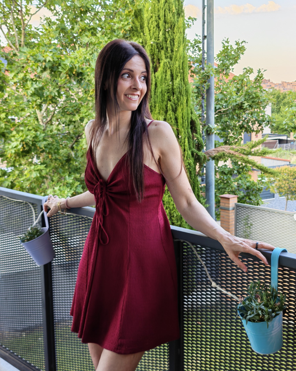 Mujer con vestido rojo en balcón, rodeada de plantas, verano en el jardín.