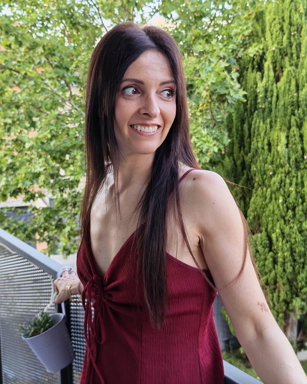Mujer sonriendo en un balcón con vestido rojo, rodeada de naturaleza verde.