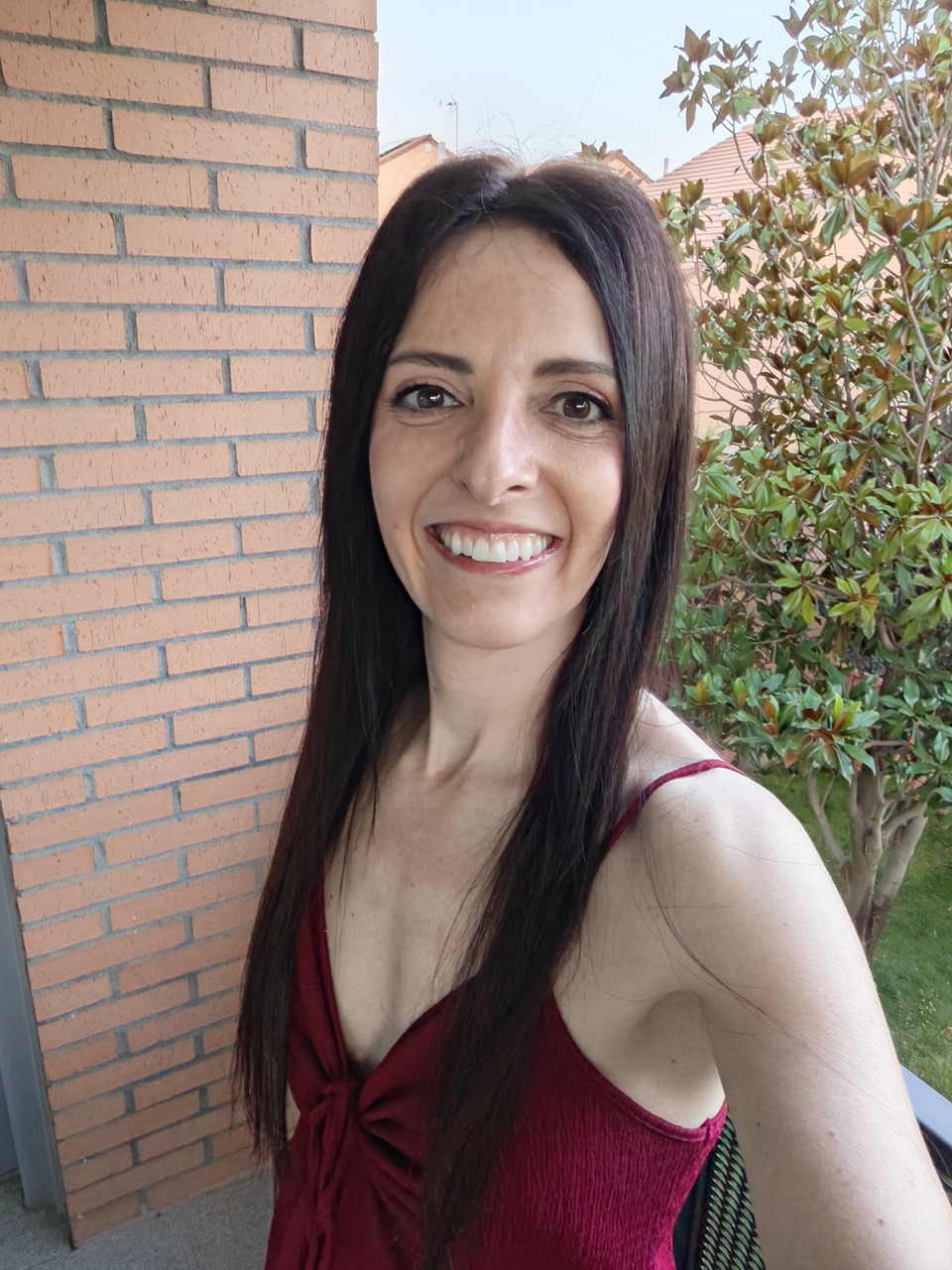 Mujer sonriendo en una terraza con vestido rojo y fondo de ladrillo y plantas, selfie al aire libre.