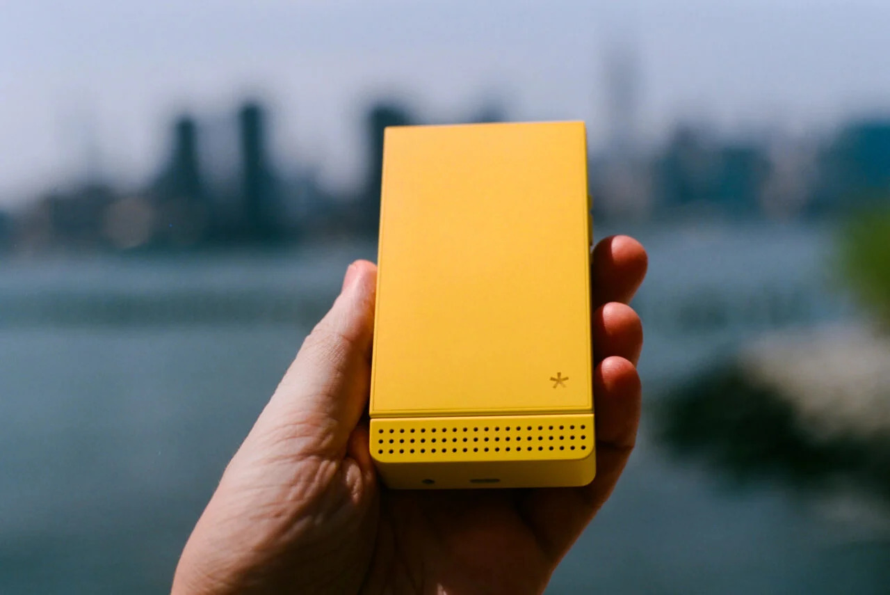 Dispositivo electrónico amarillo en mano, fondo difuminado de ciudad y agua. Tecnología portátil innovadora.