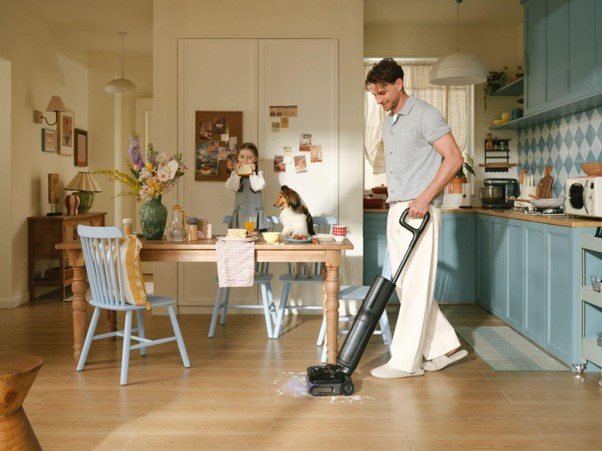 Hombre aspirando el suelo de cocina mientras niña y perro están en la mesa. Limpieza del hogar con aspiradora moderna.