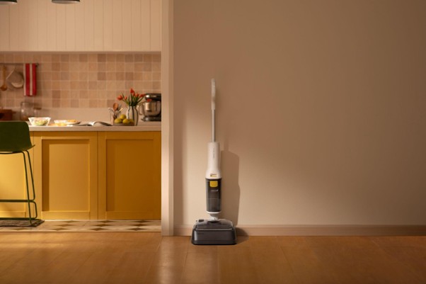Aspiradora vertical blanca apoyada en la pared de una cocina moderna con muebles amarillos y utensilios de cocina.