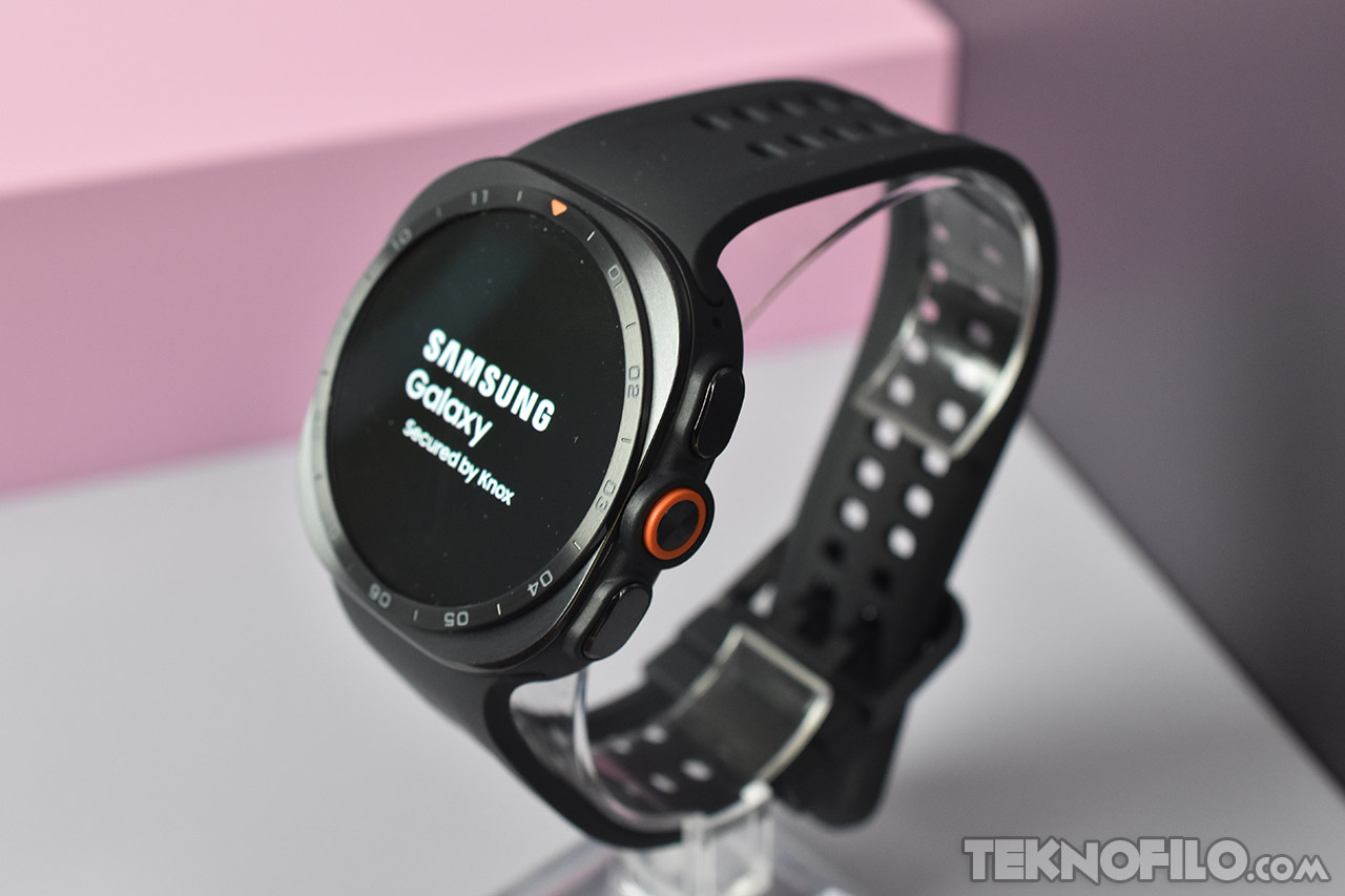 Reloj inteligente Samsung Galaxy negro con pantalla encendida, seguro con Knox. Display sobre fondo rosa y gris.