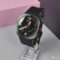 Reloj inteligente negro con pantalla analógica, sobre fondo rosa y gris. Ideal para fitness y estilo moderno.