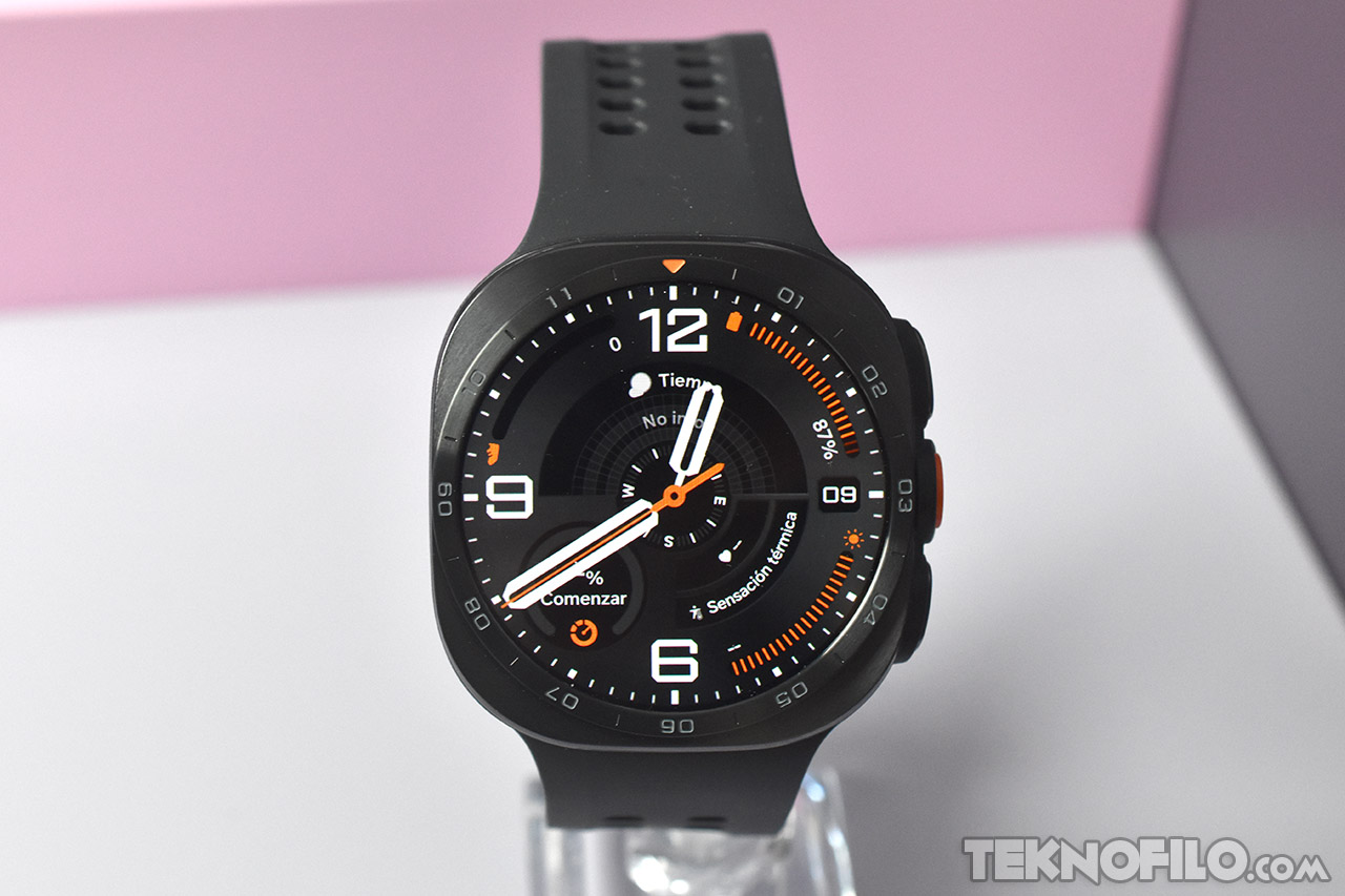 Reloj inteligente negro con pantalla digital mostrando hora, clima y otros indicadores sobre fondo rosa y gris.