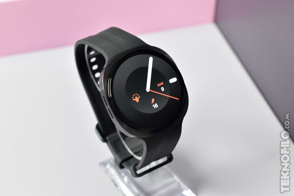 Samsung Galaxy Watch9: pantalla de 3.000 nits y Snapdragon Wear Elite Reloj inteligente negro con pantalla redonda, mostrando la fecha 9 de marzo. Correa ajustable. Fondo blanco y rosa.