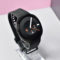 Reloj inteligente negro con pantalla redonda, mostrando la fecha 9 de marzo. Correa ajustable. Fondo blanco y rosa.