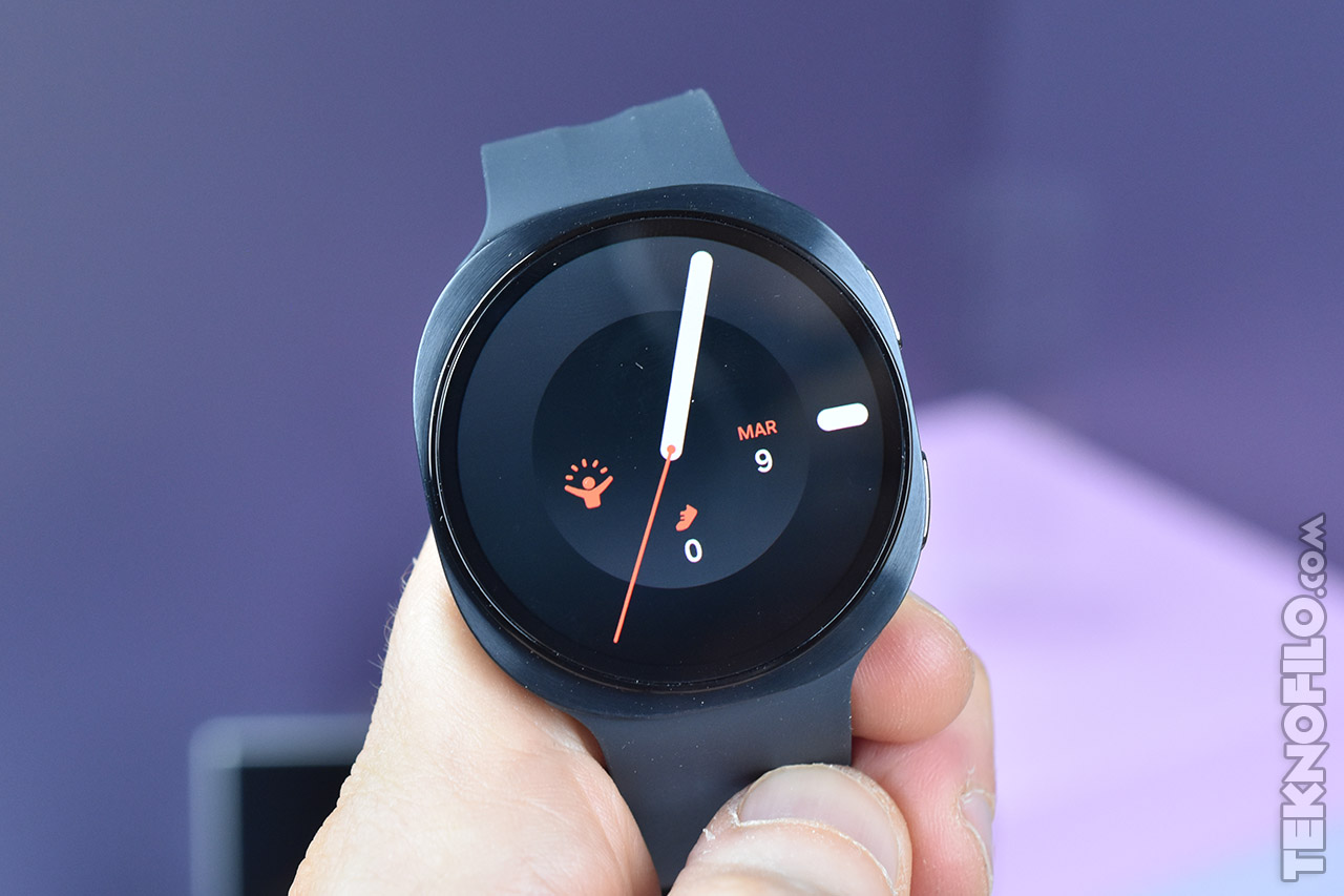 Reloj inteligente negro con pantalla digital mostrando la fecha 9 de marzo. Fondo morado difuminado.
