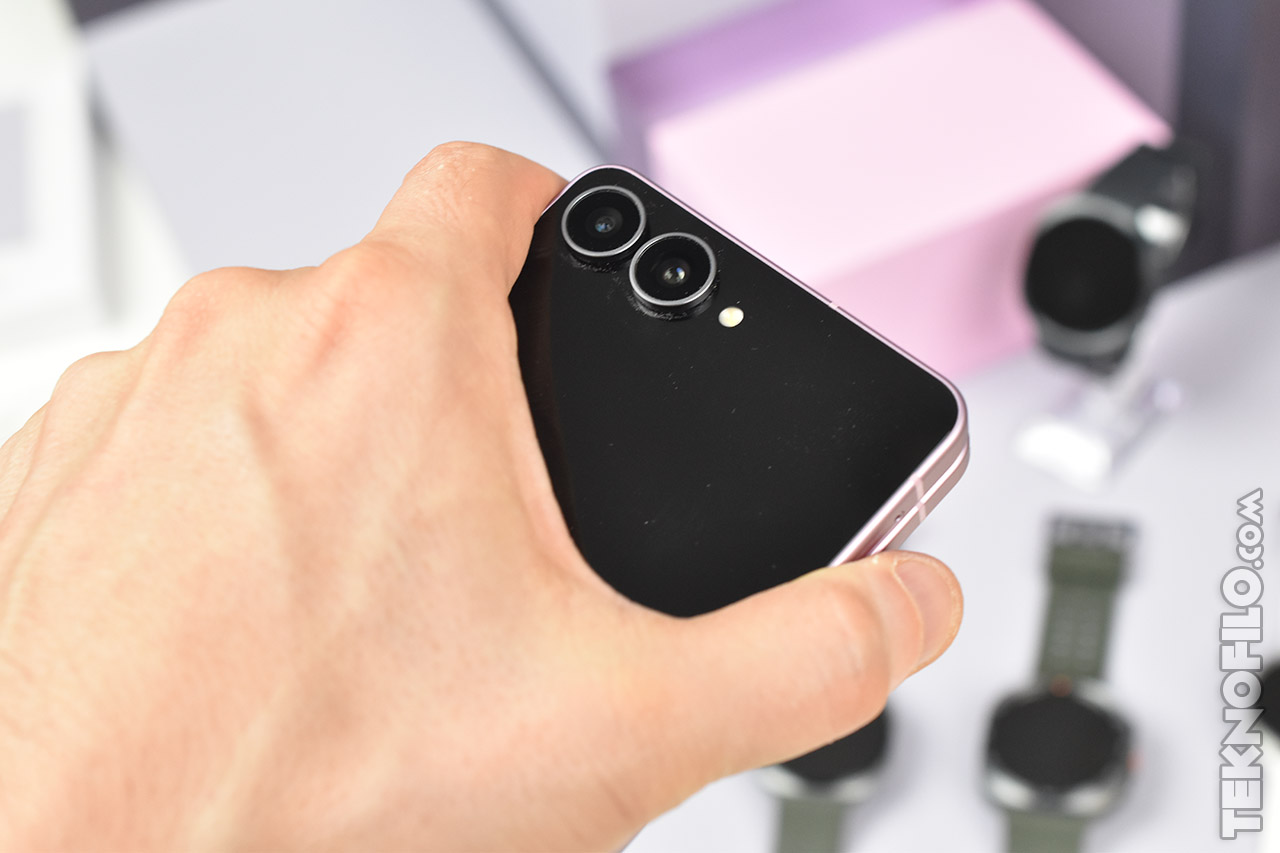 Mano sujetando un smartphone con cámara dual, fondo desenfocado de relojes y caja rosa.