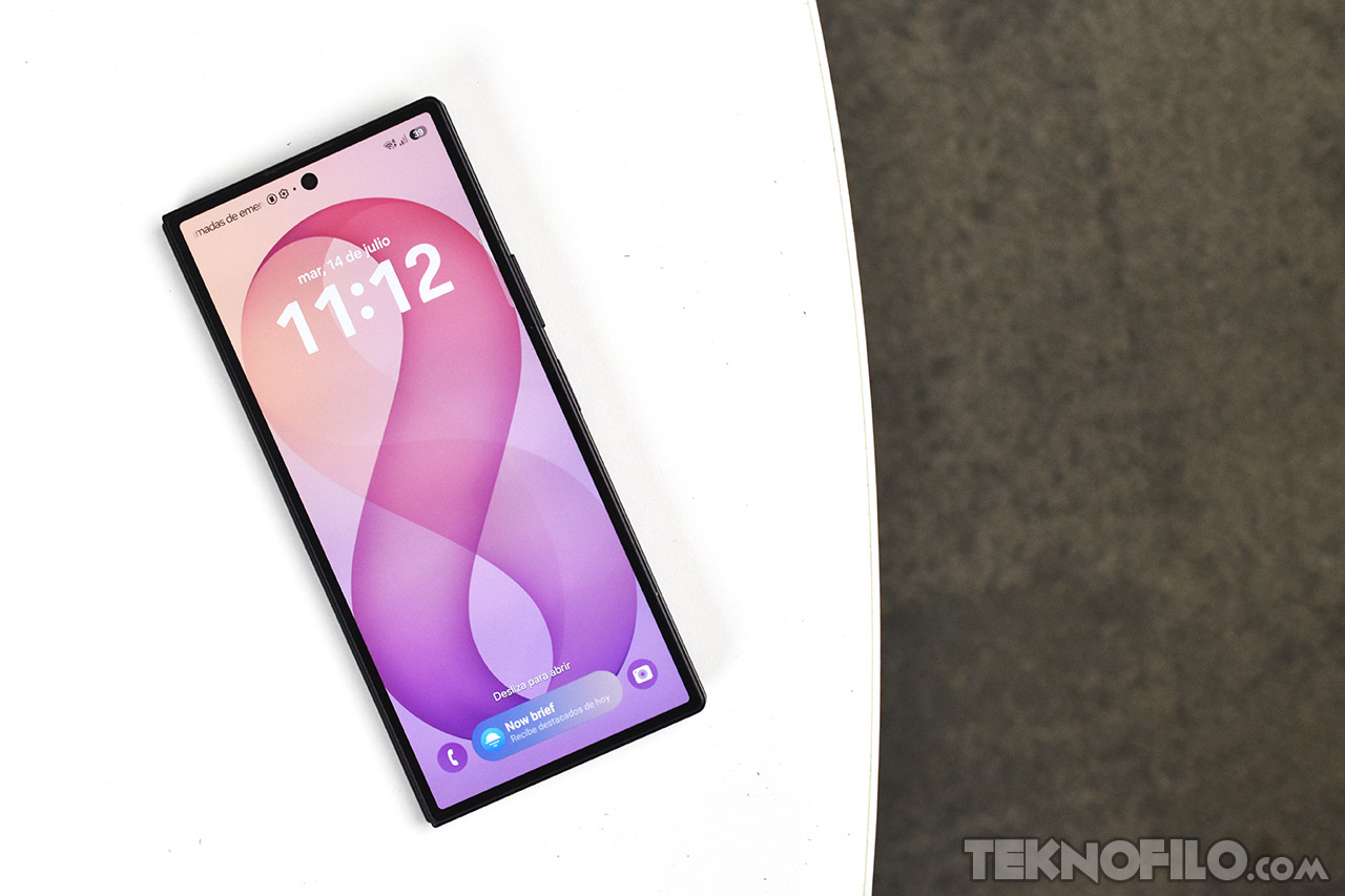 Smartphone moderno en superficie blanca, pantalla mostrando hora 11:12 y fecha 14 de julio. Diseño elegante y minimalista.