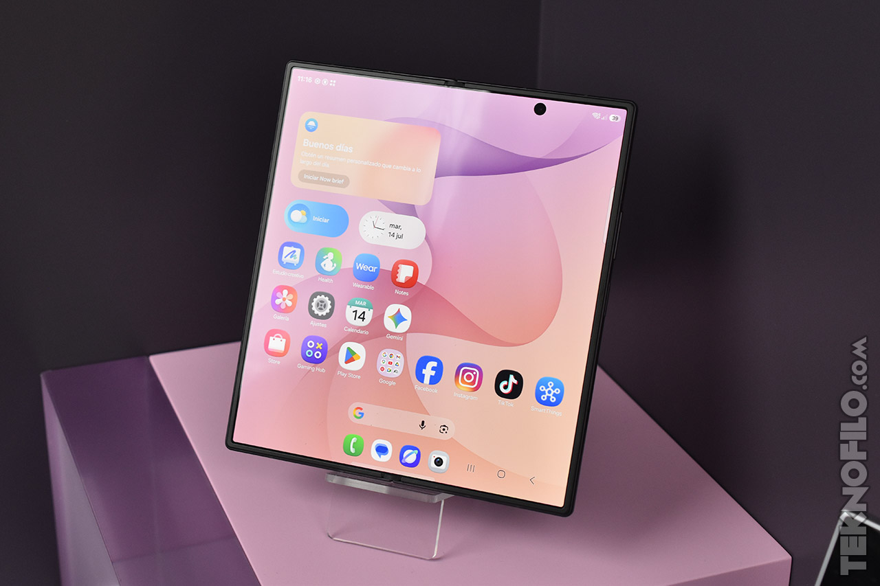 Pantalla de smartphone plegable con aplicaciones visibles sobre fondo rosa, en exhibición sobre una base violeta.