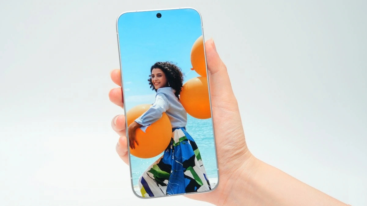 Smartphone en mano mostrando a mujer con globos naranjas en la playa, cielo azul de fondo.