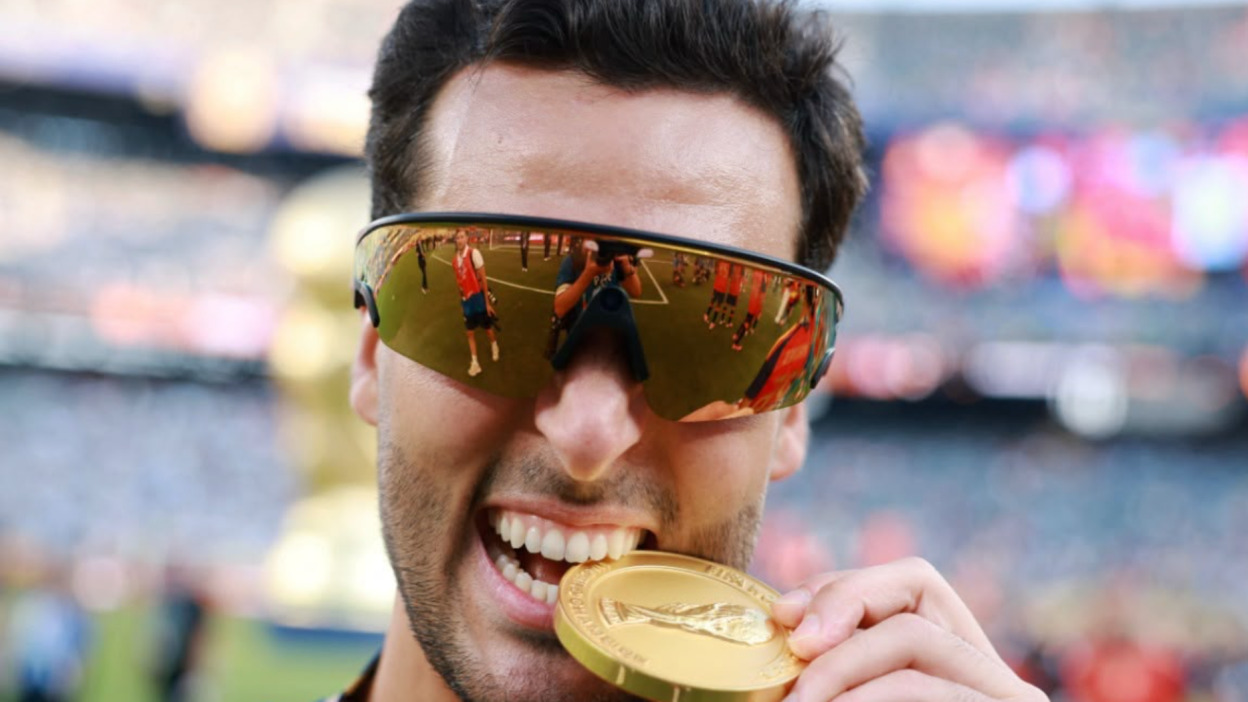 Hombre con gafas de sol mordiendo medalla de oro, reflejo de equipo de fútbol, celebración en estadio.