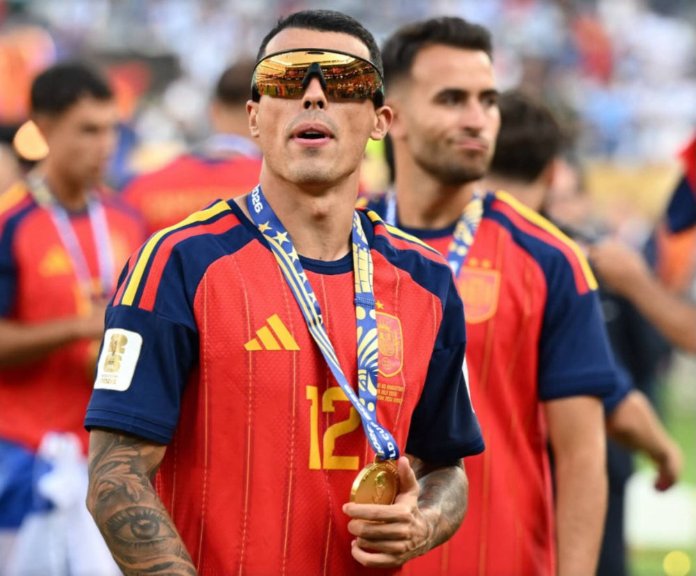Jugador de fútbol de España con medalla y gafas de sol, vestido con uniforme rojo, celebrando en el estadio.