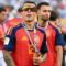 Jugador de fútbol de España con medalla y gafas de sol, vestido con uniforme rojo, celebrando en el estadio.