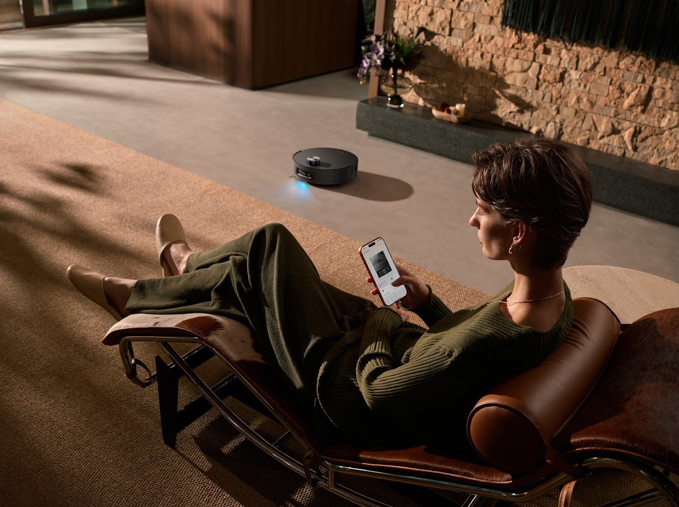 Persona relajada controla robot aspirador con smartphone desde sofá moderno. Limpieza inteligente en hogar cómodo.