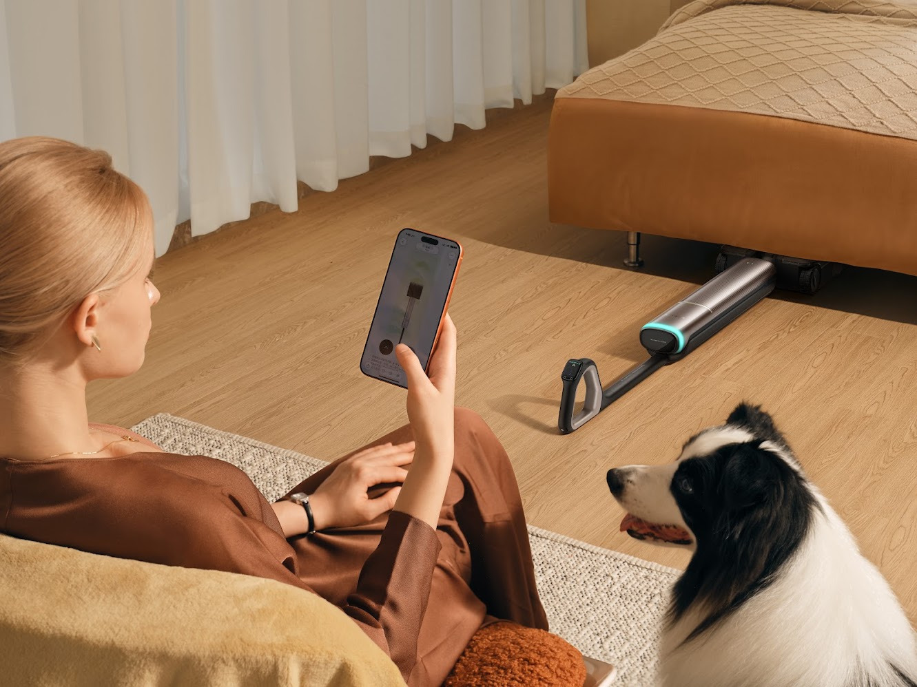 Mujer controlando robot aspirador con smartphone mientras perro observa, en sala moderna con suelo de madera.