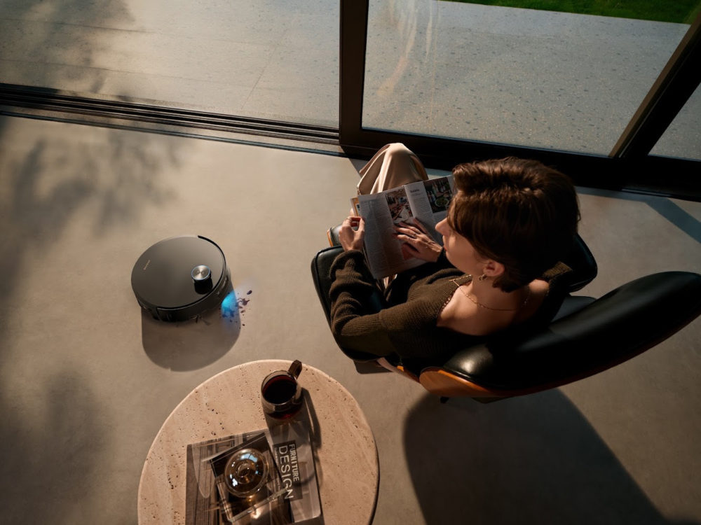 Dreame presenta en IFA 2026 un robot con vapor, cámara 8K, cortacésped 4×4 y más Persona lee mientras aspiradora robot limpia suelo junto a ventana.