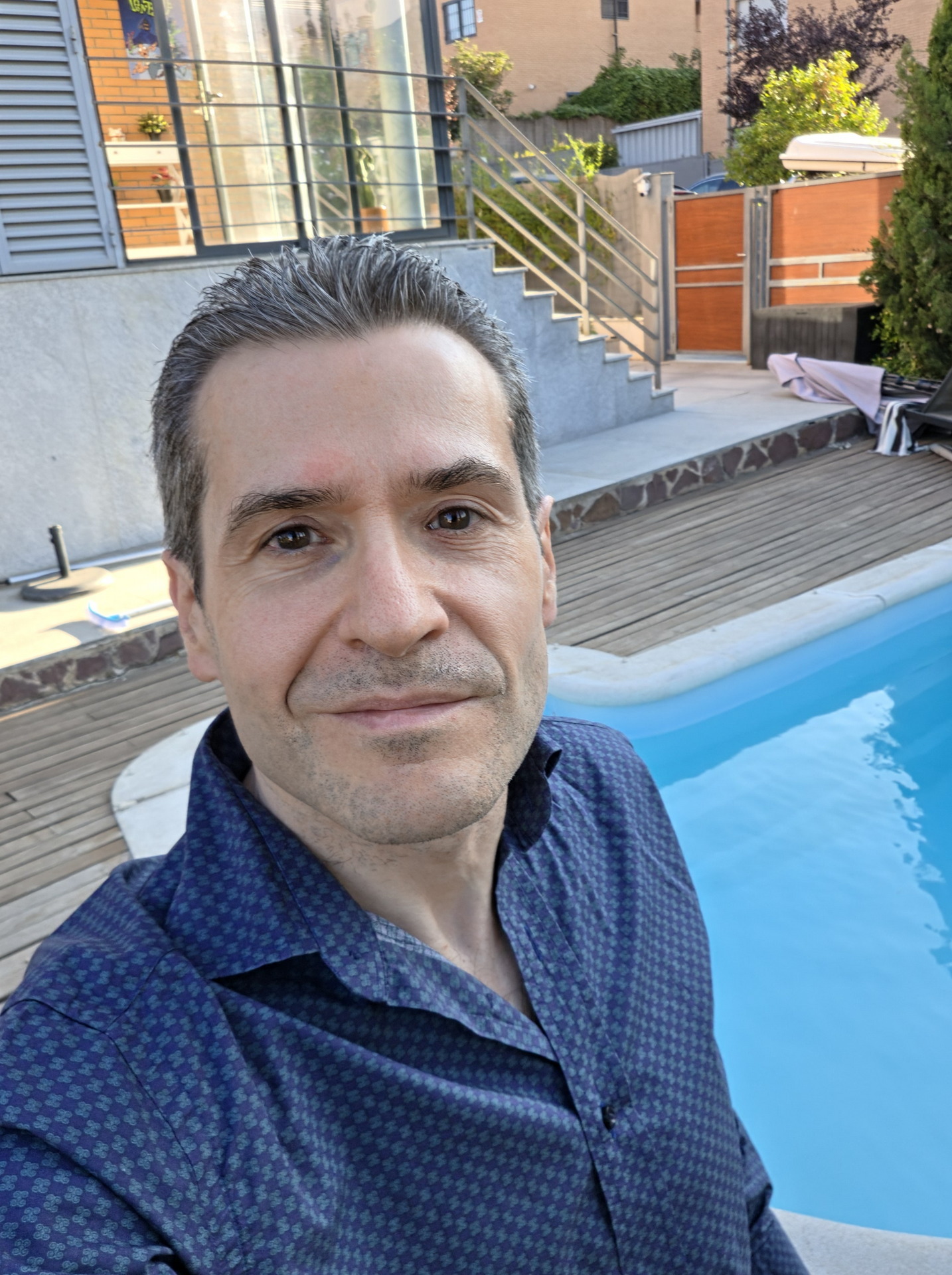 Hombre sonriendo cerca de una piscina en un patio soleado con fondo de escalera de azulejos y plantas.