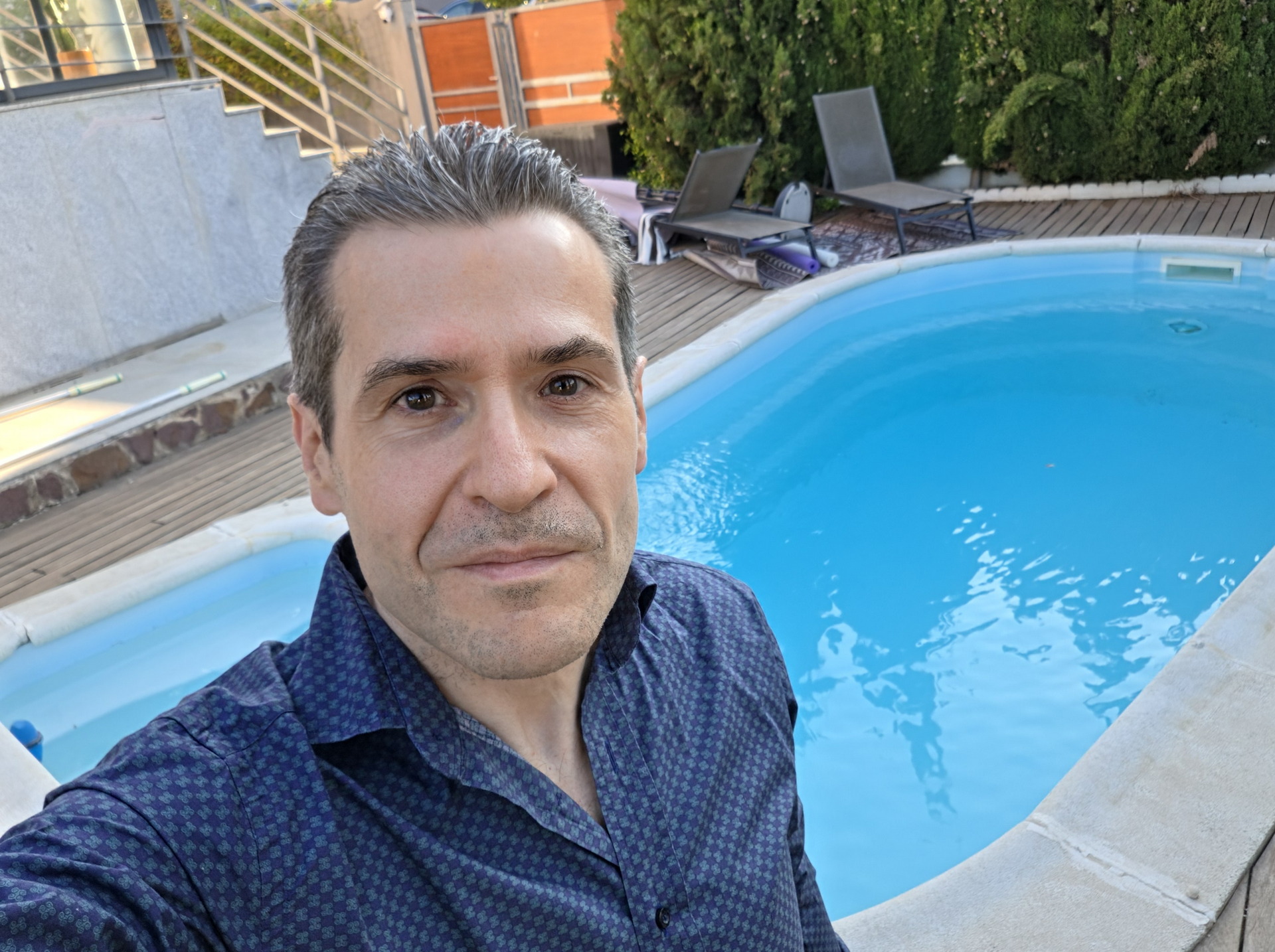 Hombre con camisa azul junto a piscina en terraza, rodeado de tumbonas y vegetación. Ambiente de relajación.