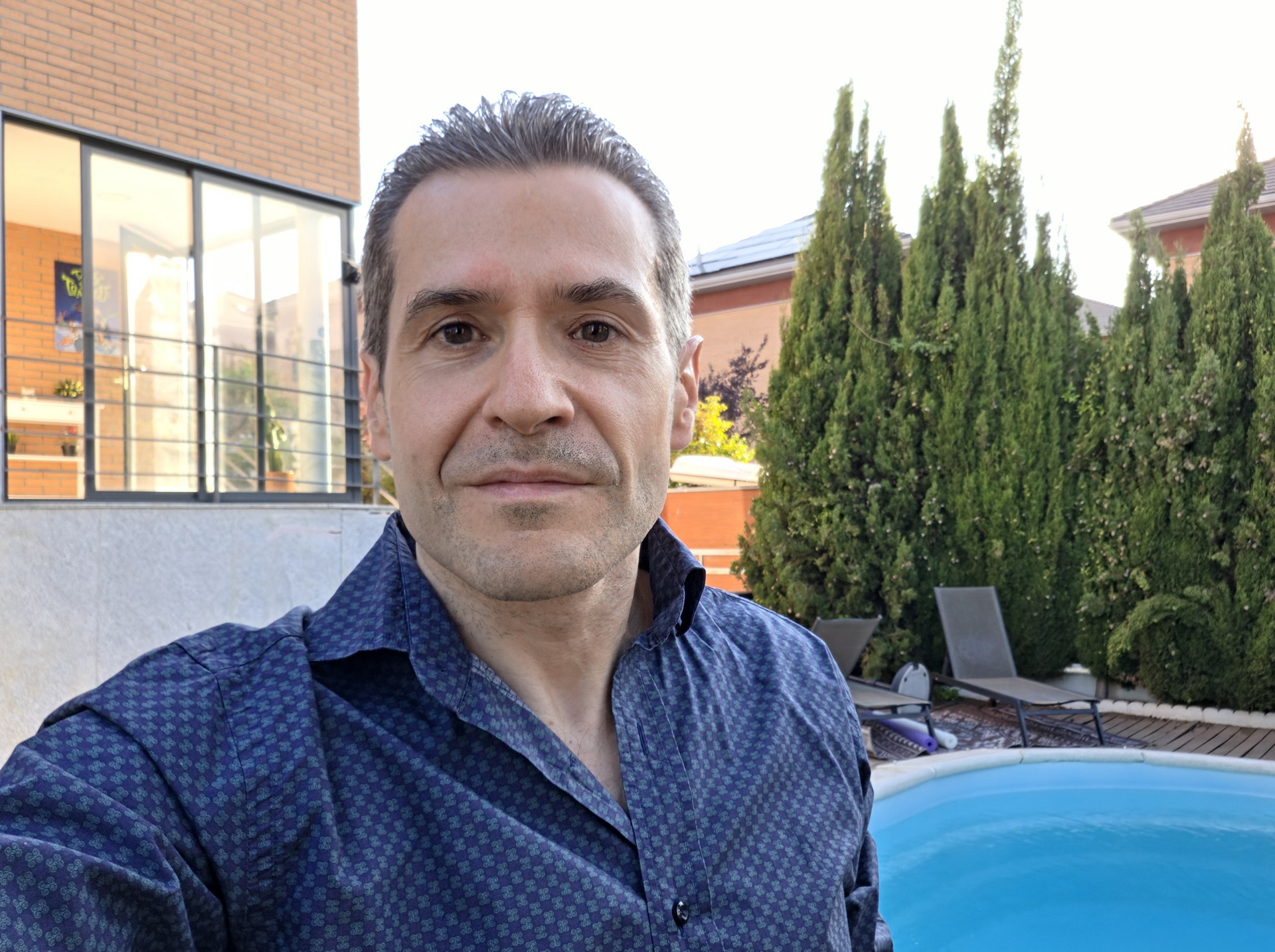 Hombre sonriendo en el jardín junto a la piscina, con fondo de casa y árboles verdes.