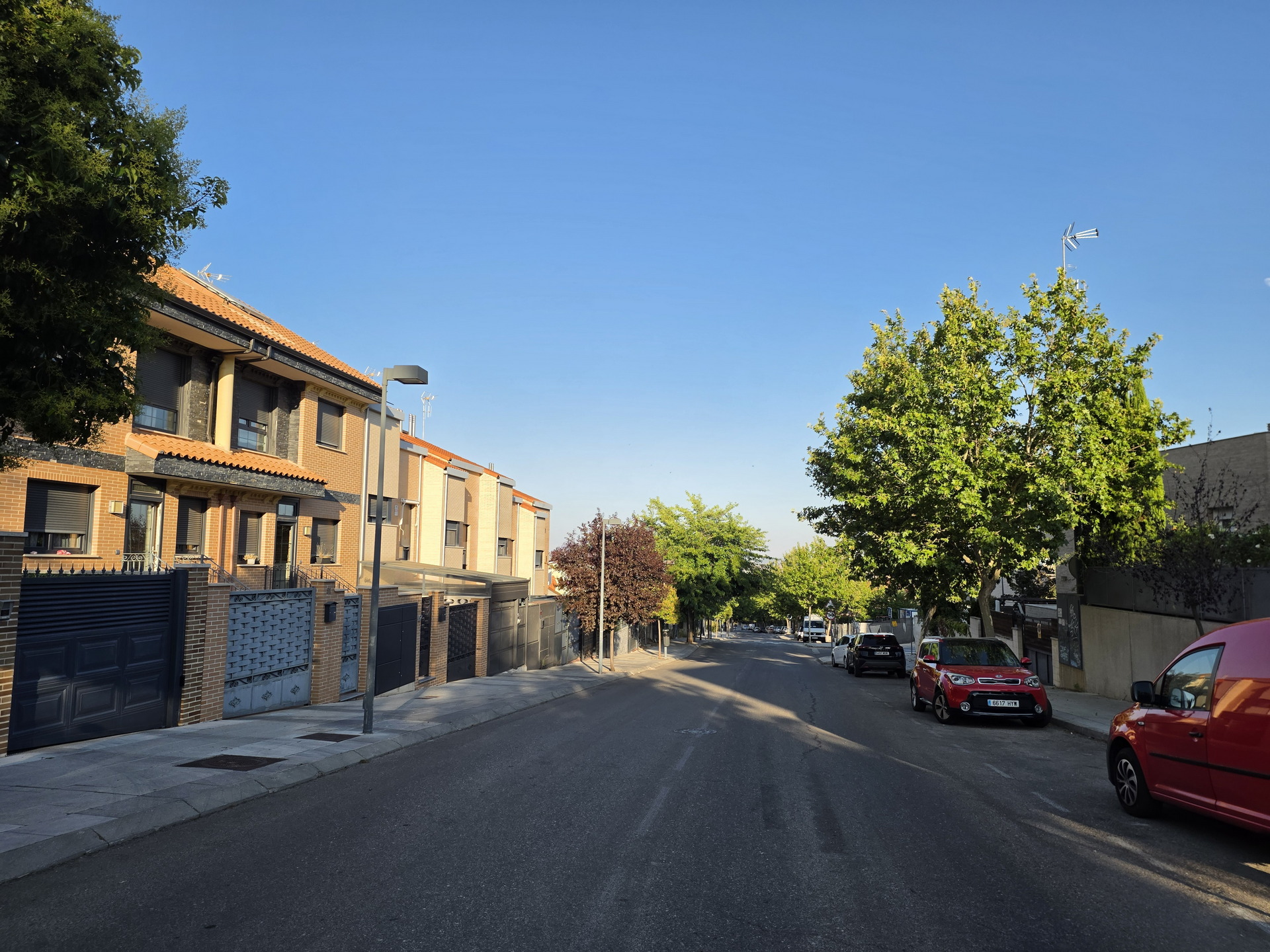 Calle residencial con casas de ladrillo, árboles verdes y coches aparcados bajo un cielo azul despejado.