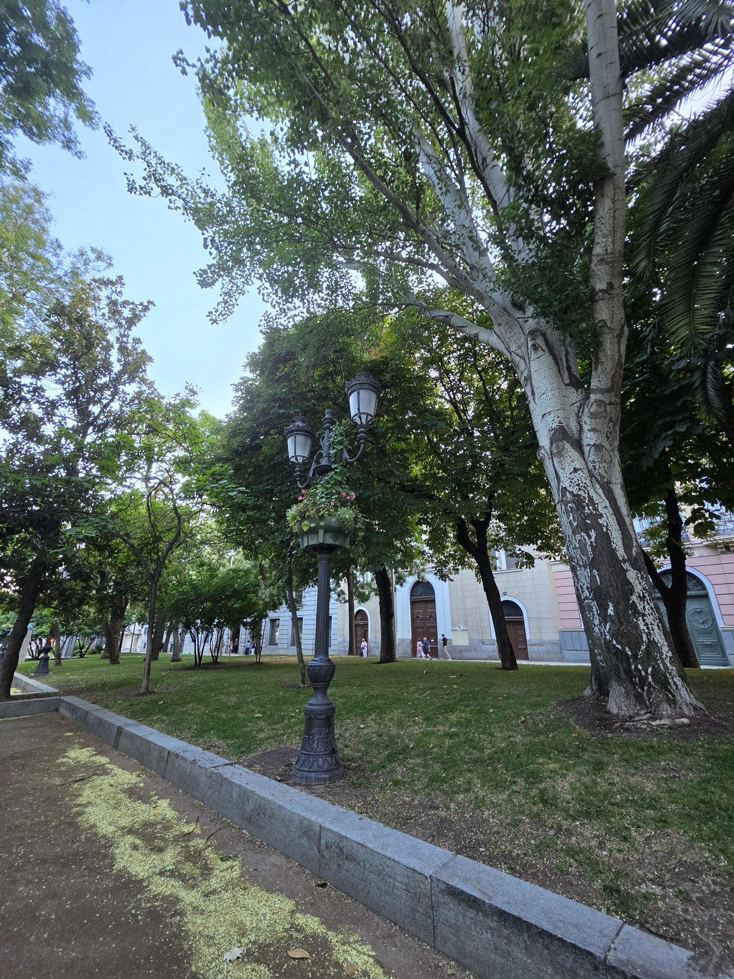 Farola clásica en parque verde con árboles altos en día soleado, cerca de edificio histórico.