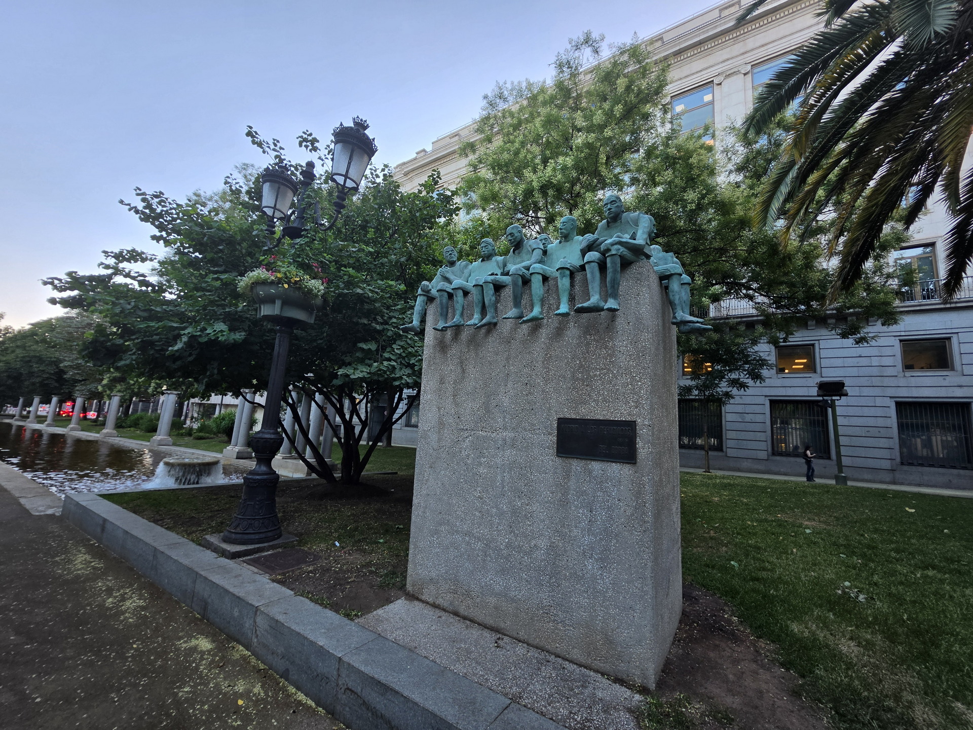 Escultura de niños sentados en un parque urbano, con edificio histórico y fuente al fondo, al atardecer.