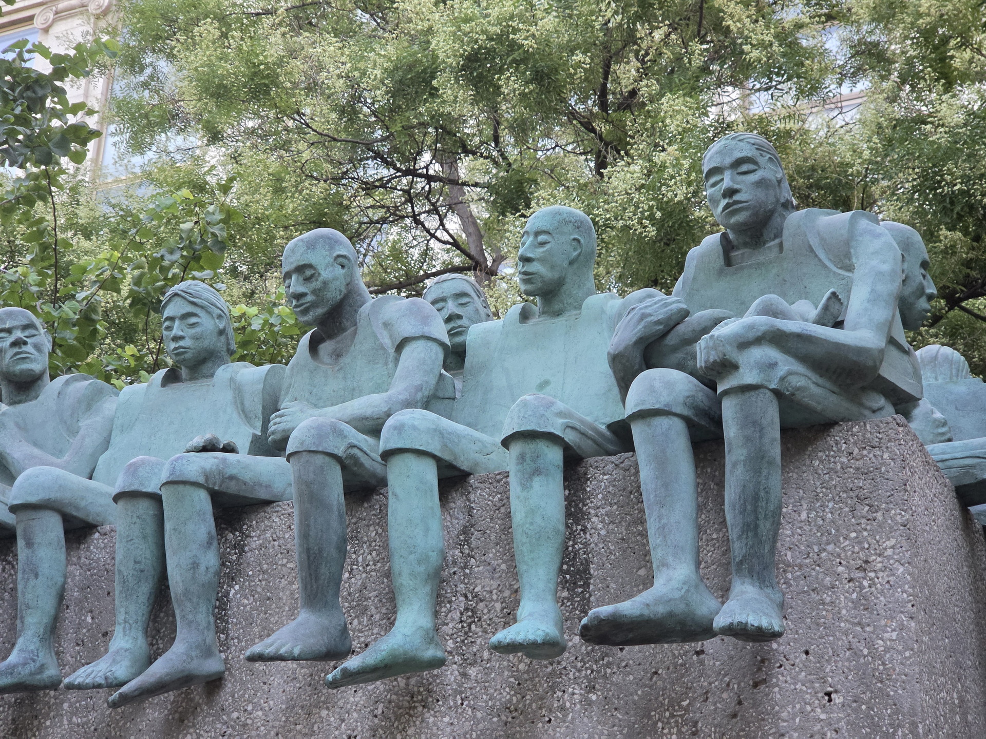 Escultura de figuras humanas sentadas de bronce en un parque, rodeadas de árboles verdes en un día soleado.