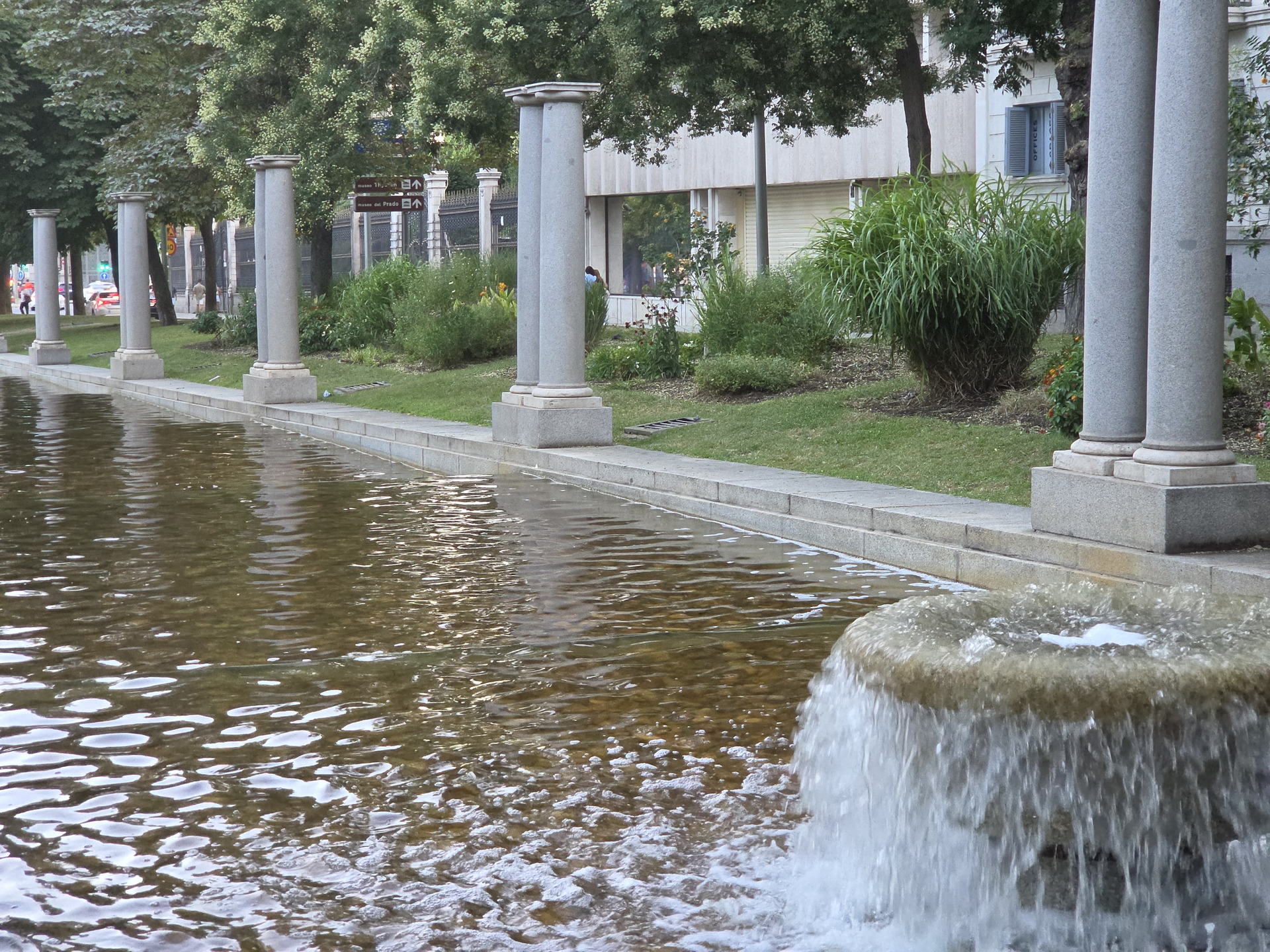 Fuente con columnas en un parque urbano rodeado de árboles y césped bien cuidado.