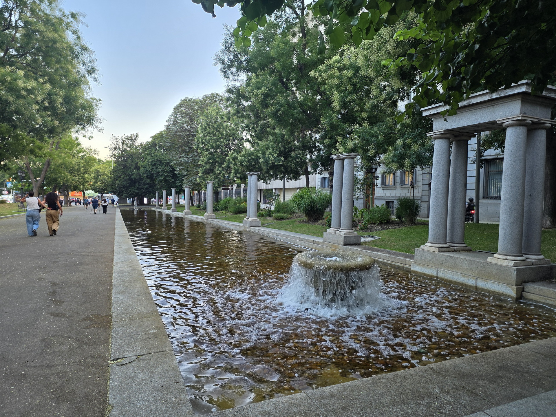 Fuente y columnas en un paseo arbolado, con personas caminando bajo un cielo despejado.