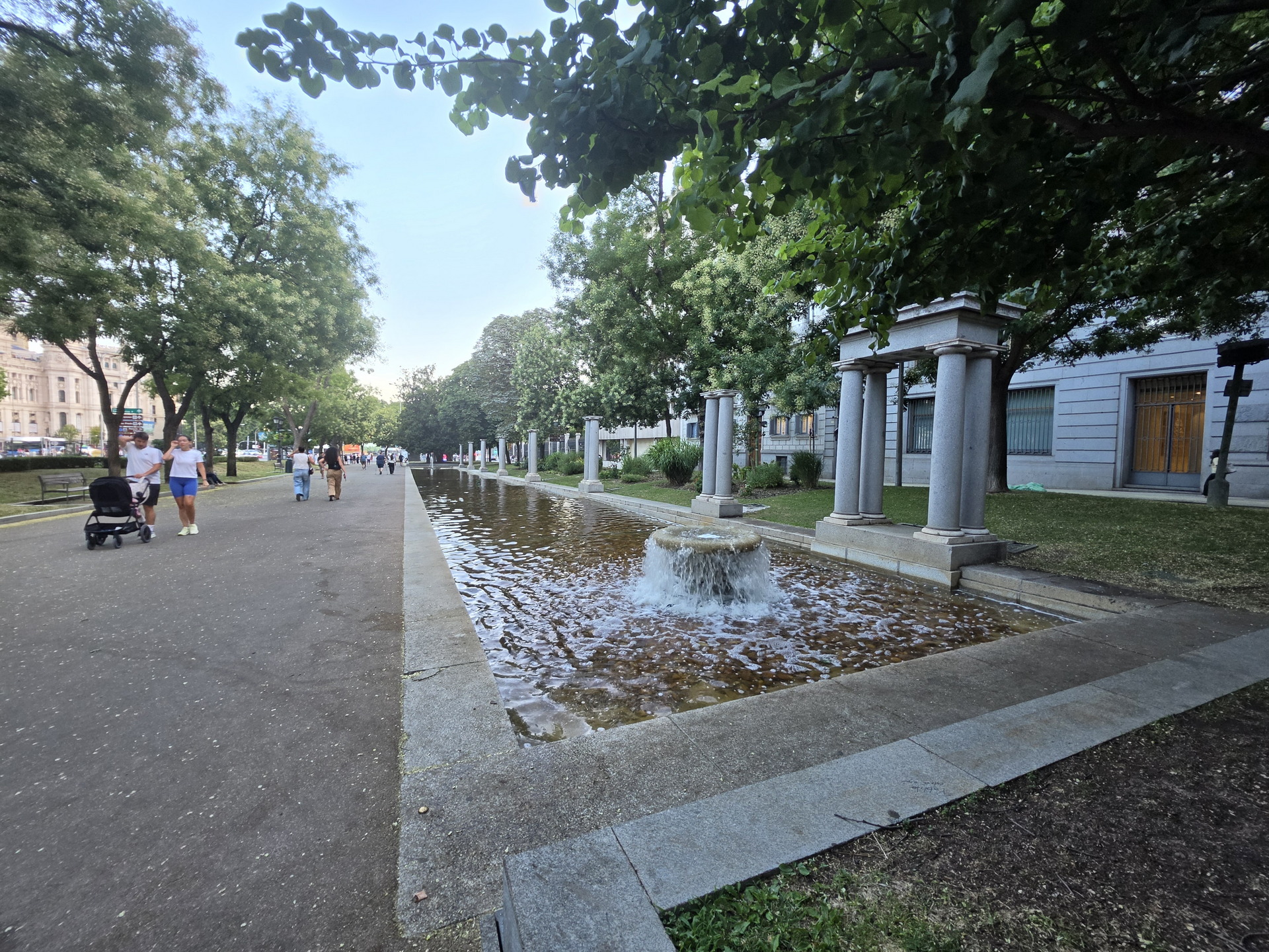 Paseo urbano con fuente, árboles y columnas clásicas, personas caminando en día soleado.