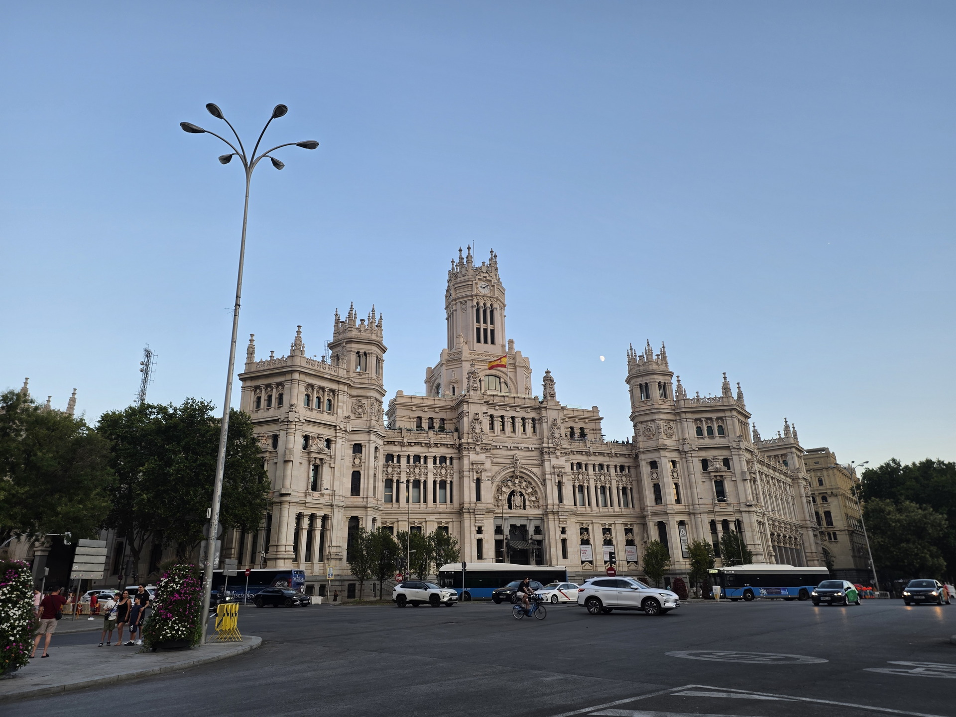 Edificio del Palacio de Cibeles en Madrid al atardecer, con coches y ciclistas circulando por el frente.