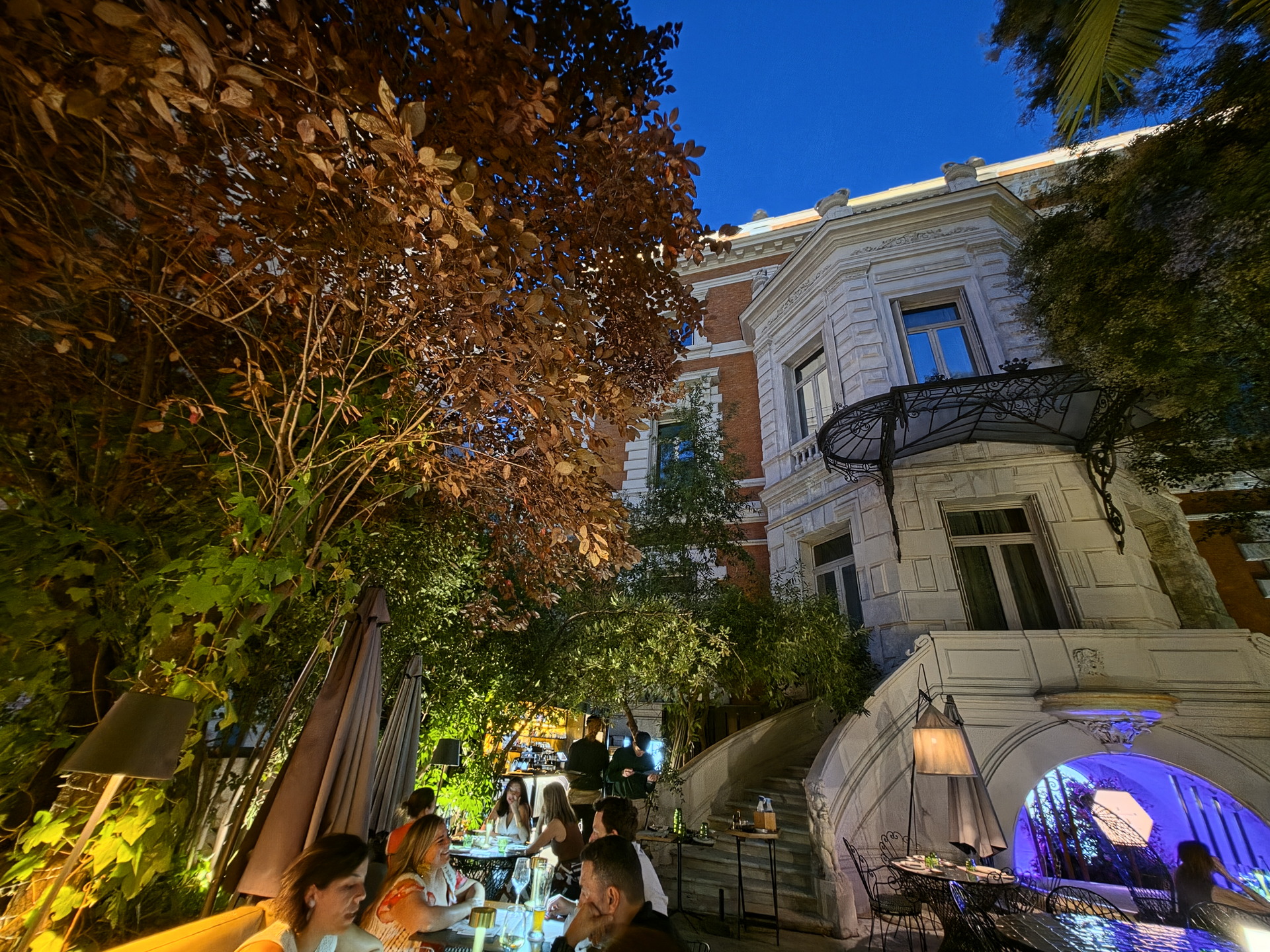 Terraza de un restaurante al aire libre con clientes, rodeada de vegetación y un edificio histórico al anochecer.
