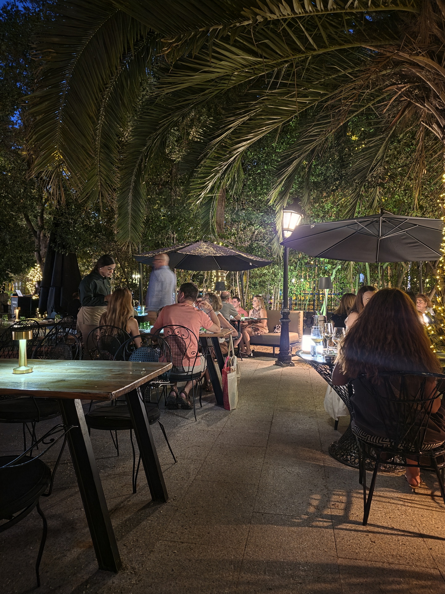 Terraza de restaurante al aire libre de noche, con personas disfrutando bajo luces y palmeras. Ambiente acogedor.