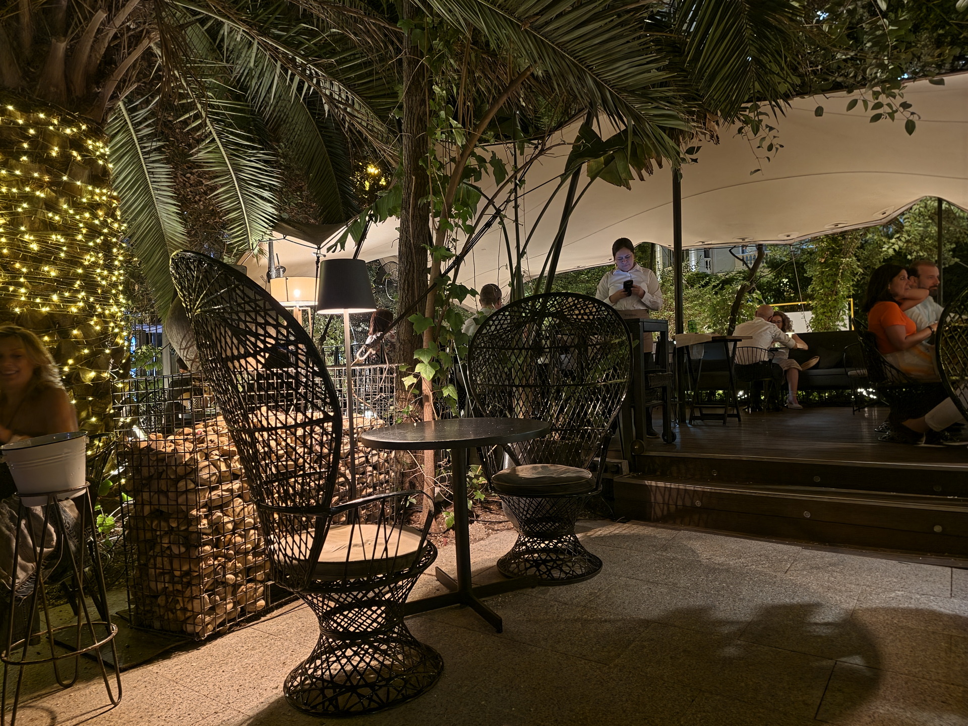 Terraza de restaurante nocturna con sillas de mimbre, luces decorativas y clientes disfrutando del ambiente.