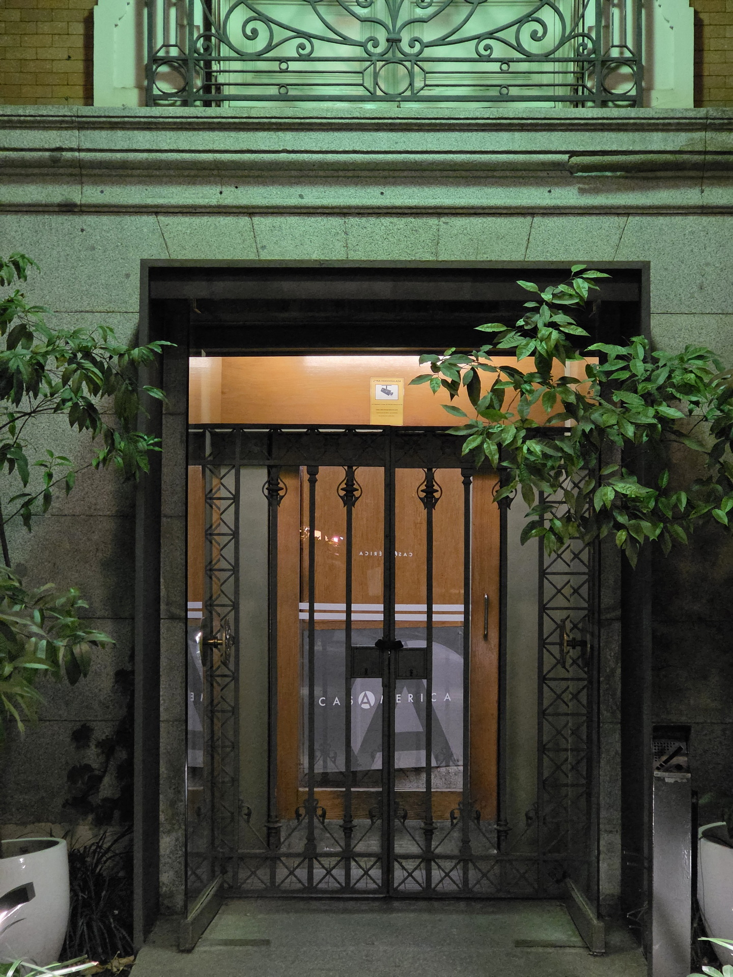 Entrada de edificio antiguo con puerta de madera y reja de hierro, decorada con plantas verdes en macetas blancas.