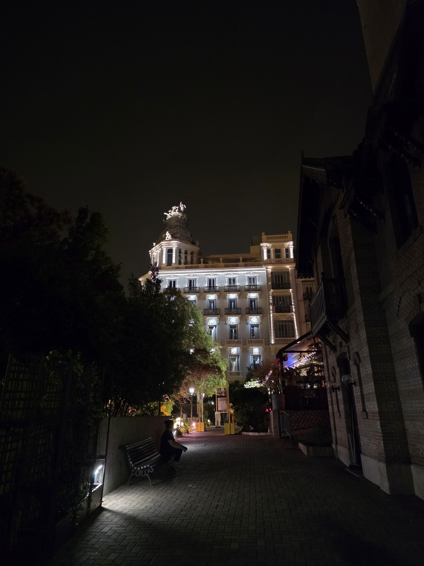Edificio iluminado de noche con estatua en la azotea, persona sentada en un banco al frente.