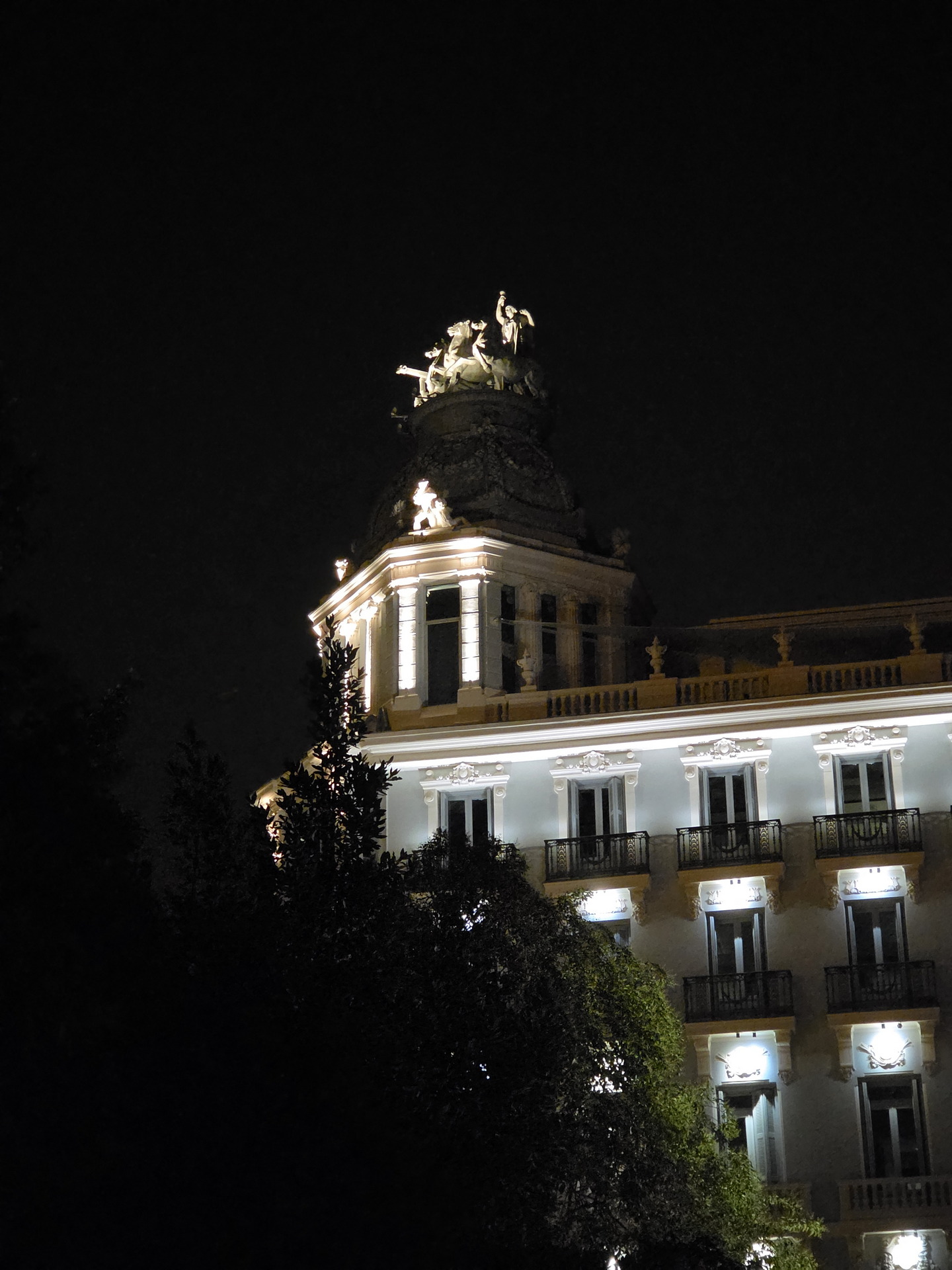 Edificio iluminado de noche con esculturas decorativas en la azotea, rodeado de árboles oscuros.