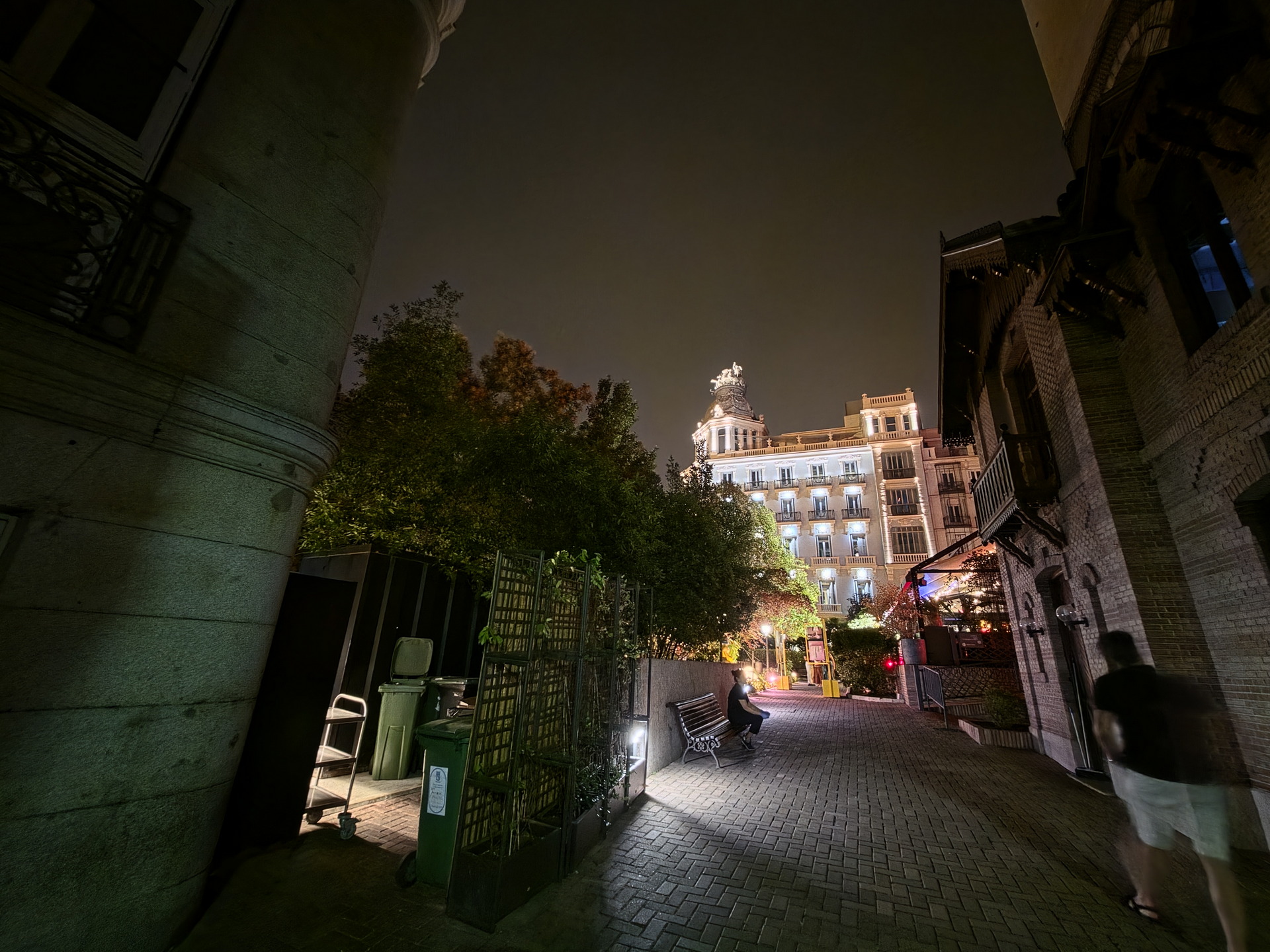 Calle iluminada por la noche con edificio histórico al fondo y árboles alrededor. Ambiente urbano y tranquilo.