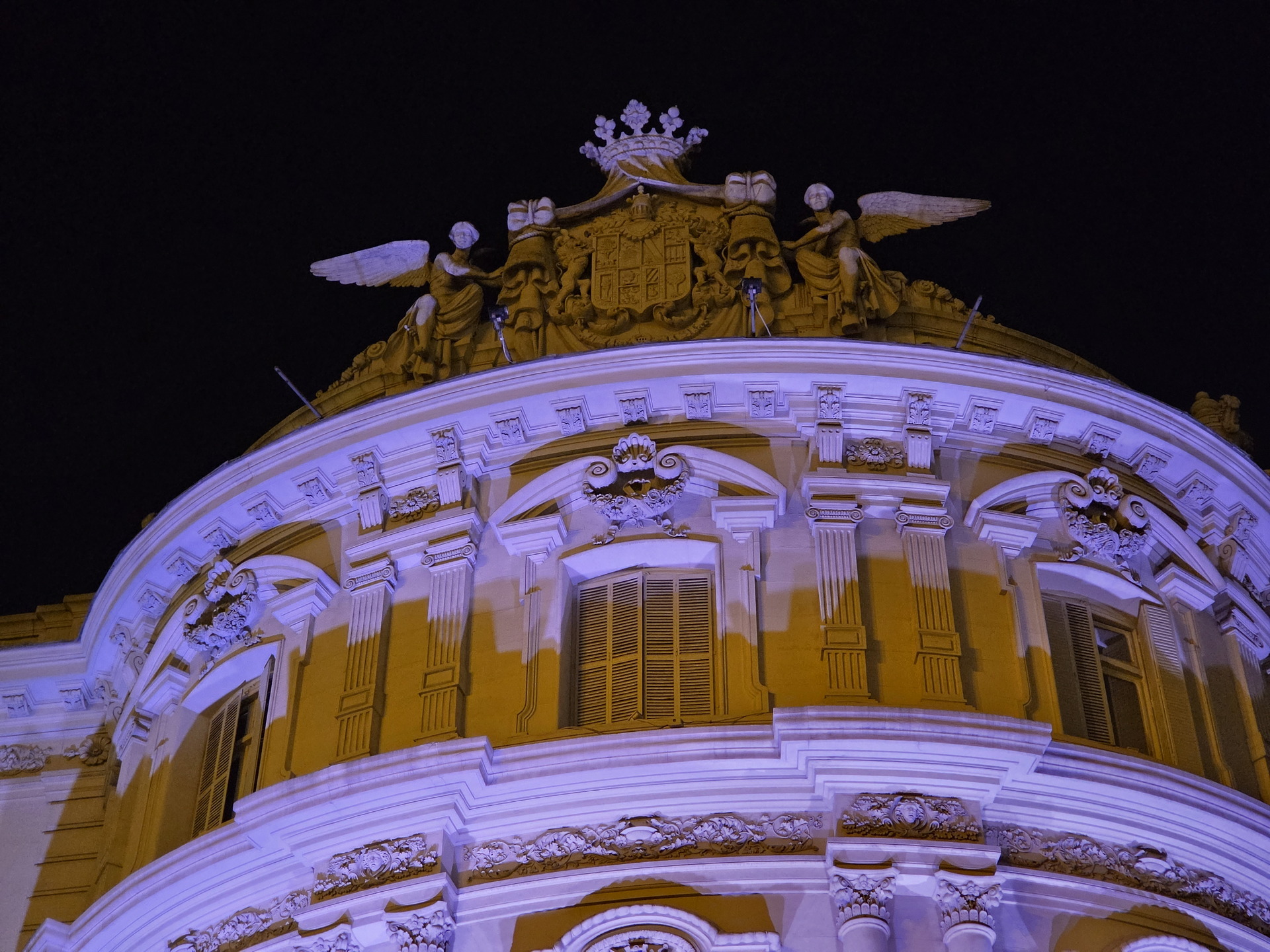 Fachada de edificio clásico iluminado de noche con esculturas y detalles arquitectónicos en tonos morados.