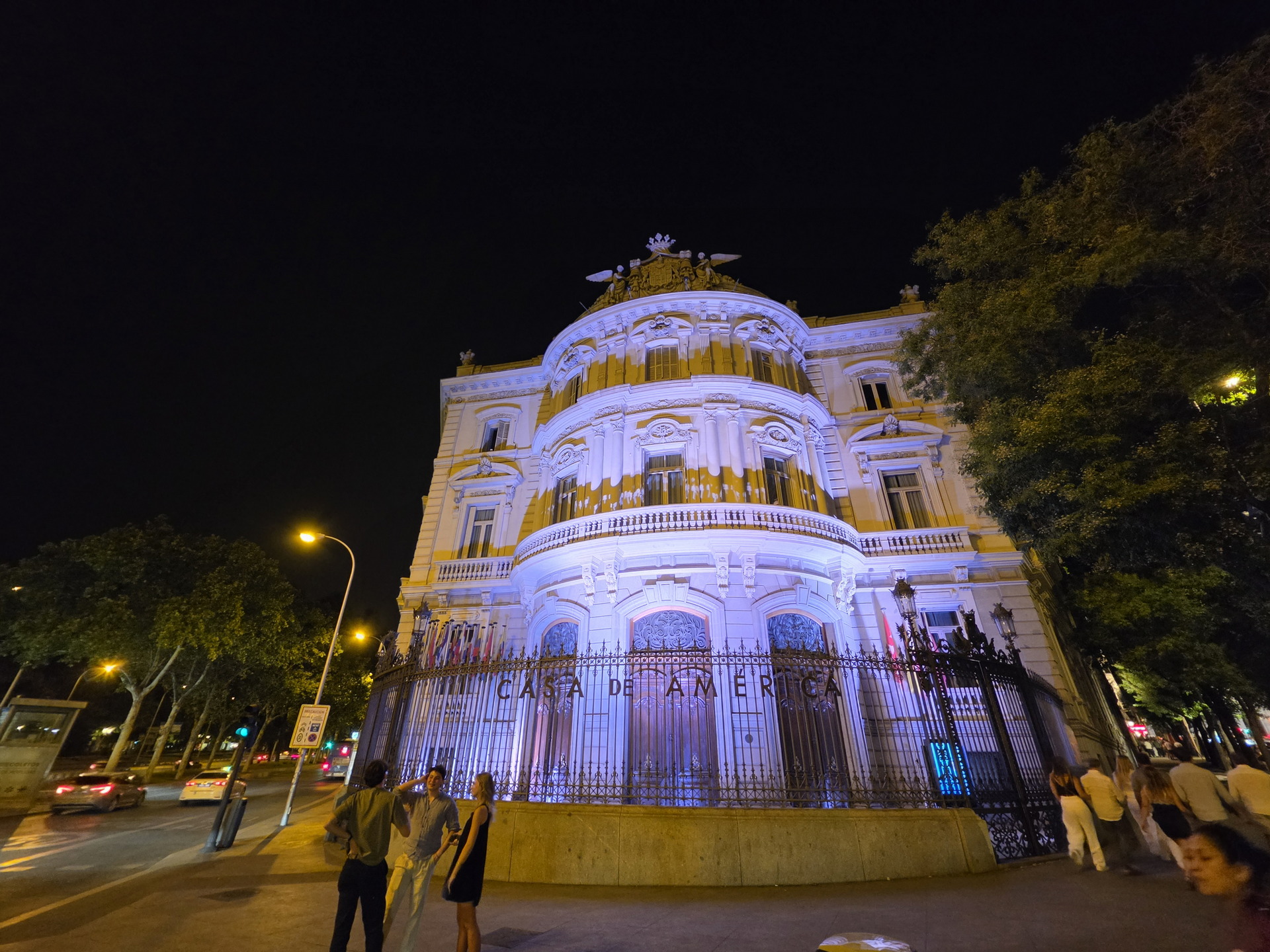 Edificio iluminado de la Casa de América en Madrid por la noche con transeúntes en primer plano.
