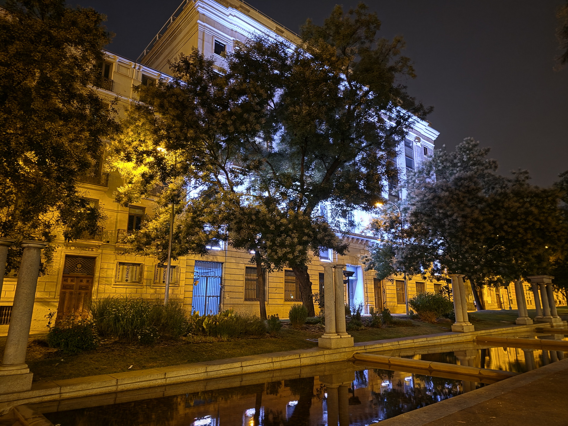 Edificio iluminado por la noche con reflejos en el agua y árboles en el entorno urbano.