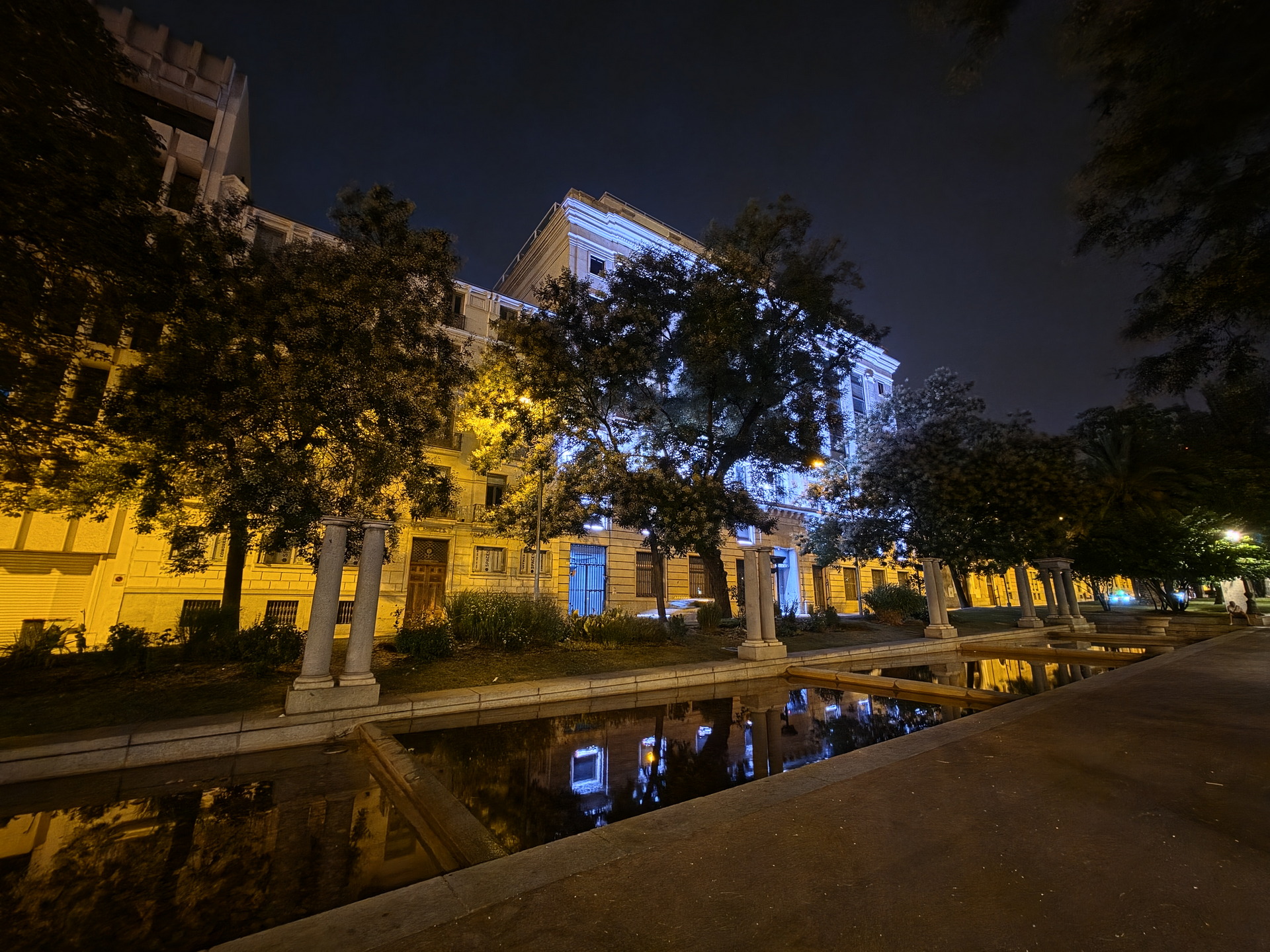 Edificio iluminado nocturno con árboles y estanque en el frente, creando una atmósfera tranquila en la ciudad.