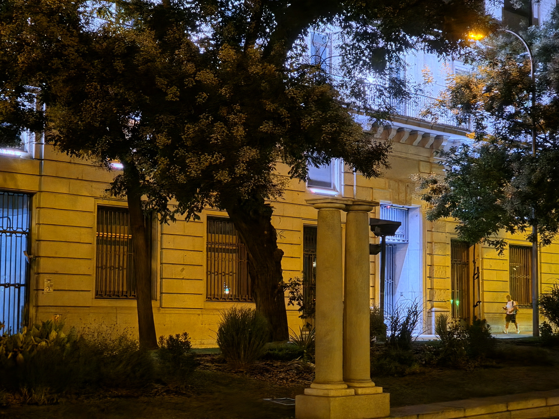 Edificio iluminado por la noche con árboles y columnas en la acera, persona caminando cerca de la entrada.