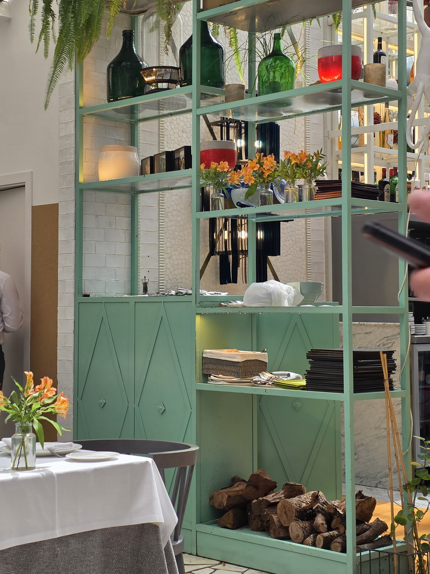 Estante verde en restaurante elegante con botellas, flores y leña. Ambiente acogedor y decorativo.