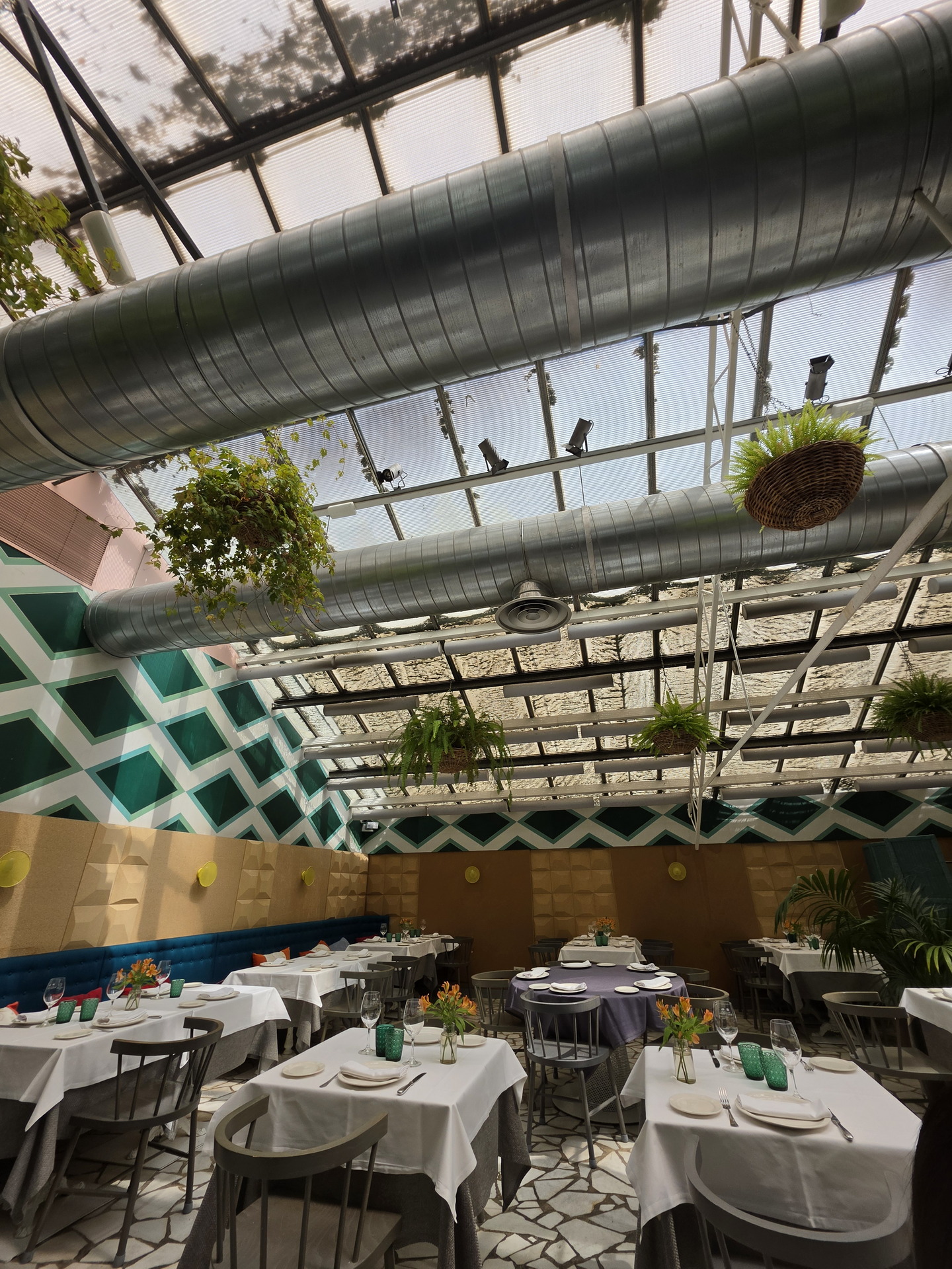 Restaurante con techo de vidrio, decoración moderna y mesas elegantes, luz natural y plantas colgantes.